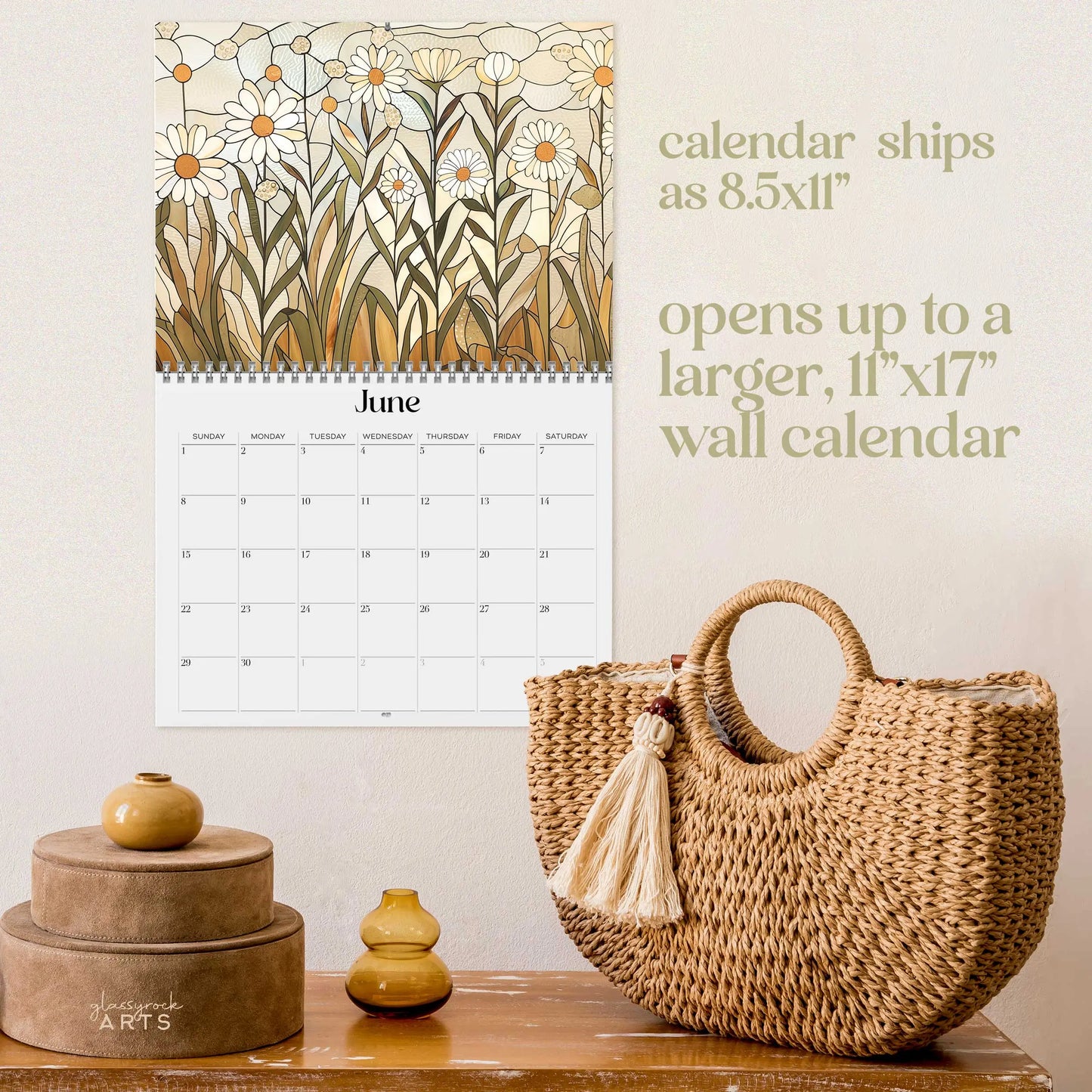 The 2025 Folk Art Flowers Wall Calendar is open to June, displaying floral art like stained glass on beige. Sizes are listed. Below rests a woven bag with a tassel, a small yellow jar, and two stacked round boxes on wood.
