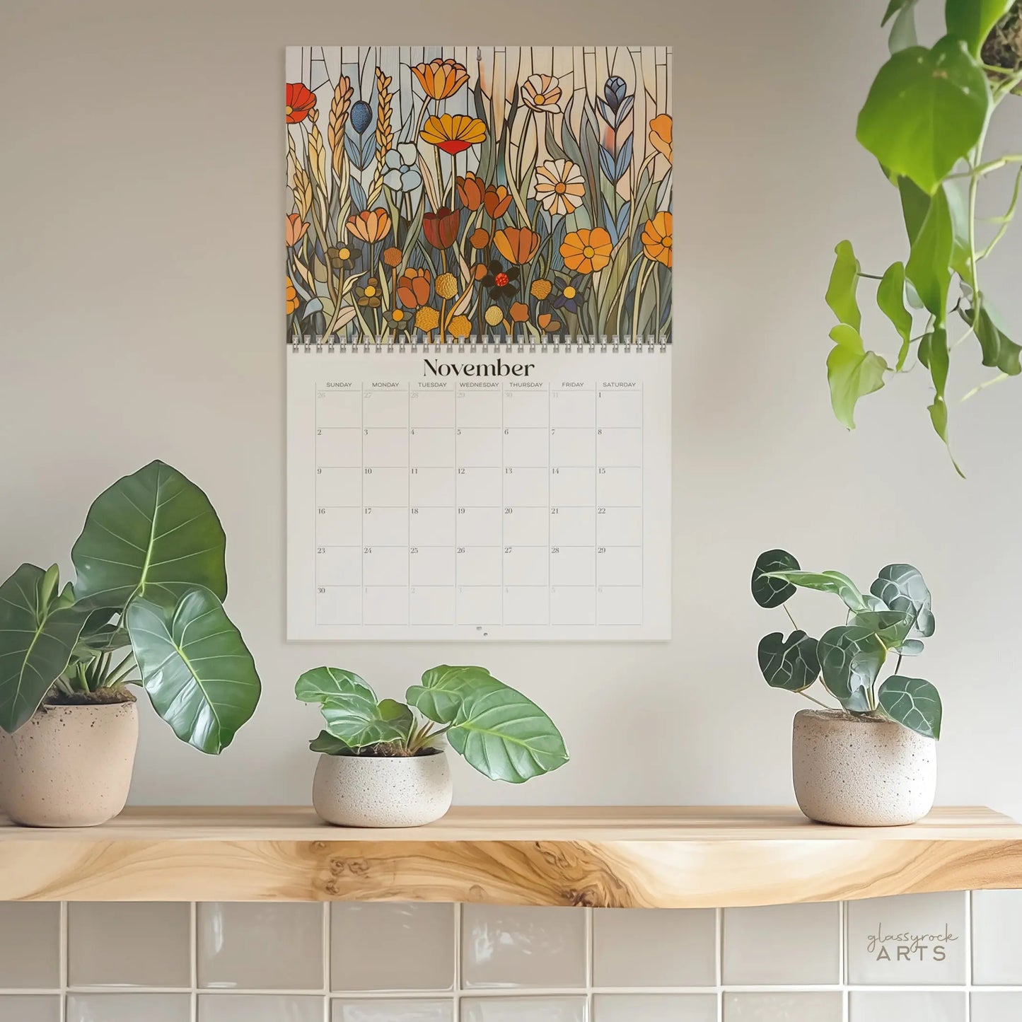 The 2025 Folk Art Flowers Wall Calendar hangs on a light-colored wall, displaying November’s page. Above the grid, an illustration bursts with folk art-inspired colorful flowers, while three potted plants sit elegantly on a wooden shelf below.