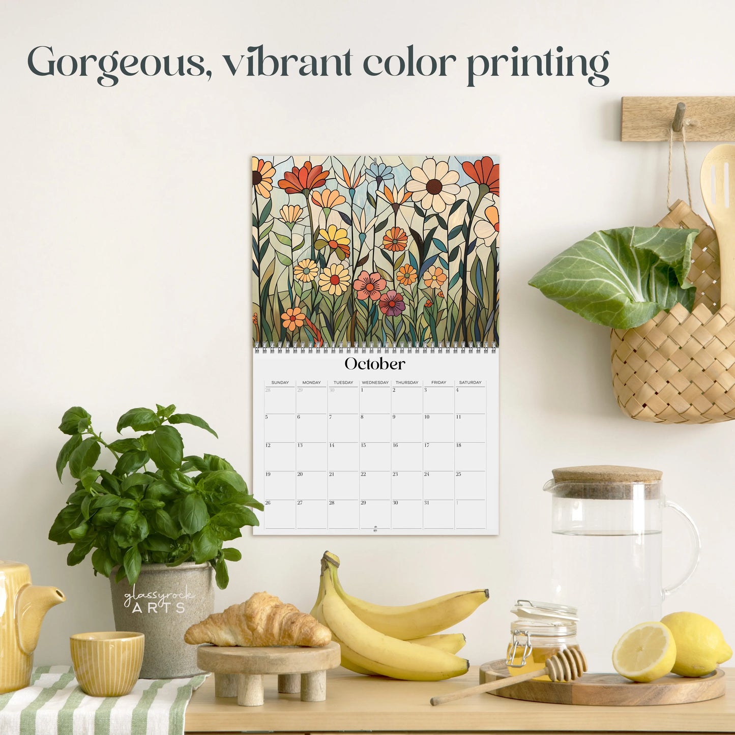 A kitchen scene featuring the 2025 Folk Art Flowers Wall Calendar open to October. Below, a counter displays bananas, a glass pitcher, a patterned potted plant, and wooden utensils in a basket. The wall text boasts Gorgeous, vibrant color printing.