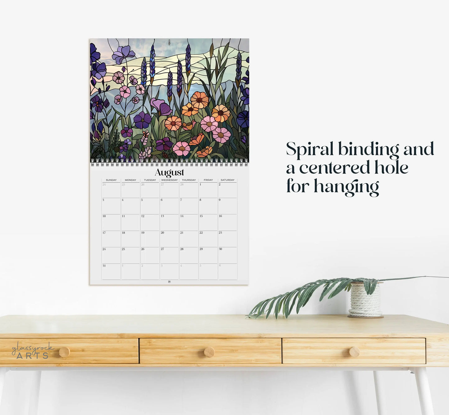 The 2025 Folk Art Flowers Wall Calendar for August displays colorful wildflower illustrations above the dates. Its spiral-bound with a center hole for hanging, beautifully set on a light wooden desk next to a leafy plant.