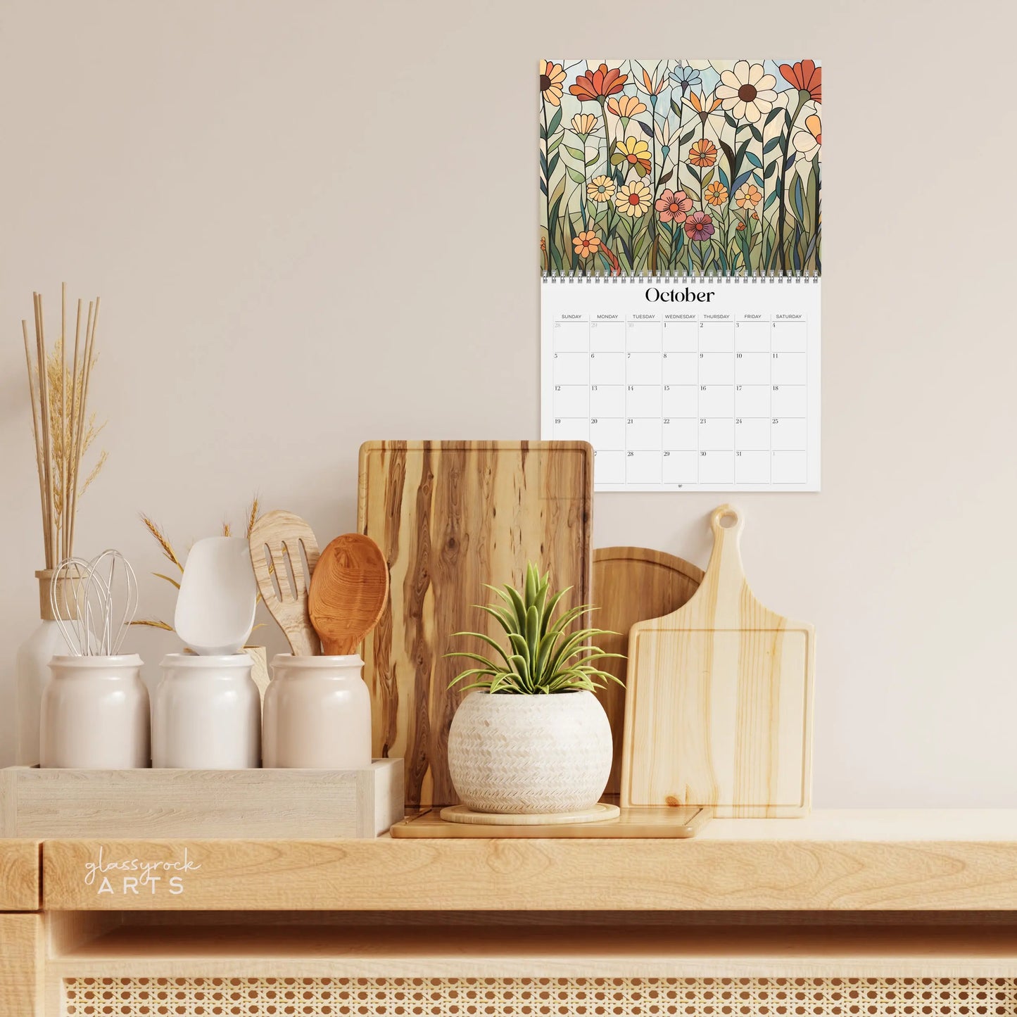 A kitchen countertop features a potted plant, three white canisters, wooden spoons, and cutting boards. Above, the 2025 Folk Art Flowers Wall Calendar is open to October, showcasing an illustrated floral design.