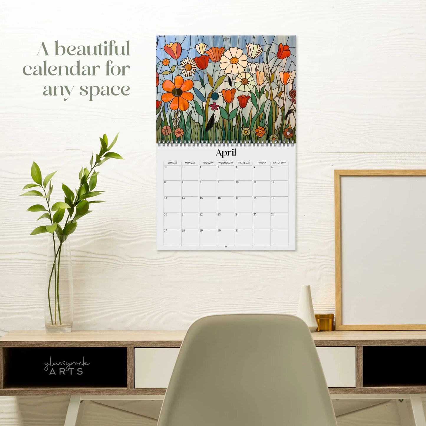The 2025 Folk Art Flowers Wall Calendar featuring a colorful floral design for April hangs on a white wall above a desk adorned with a plant, books, and decorative items. Text on the wall states, A beautiful calendar for any space.
