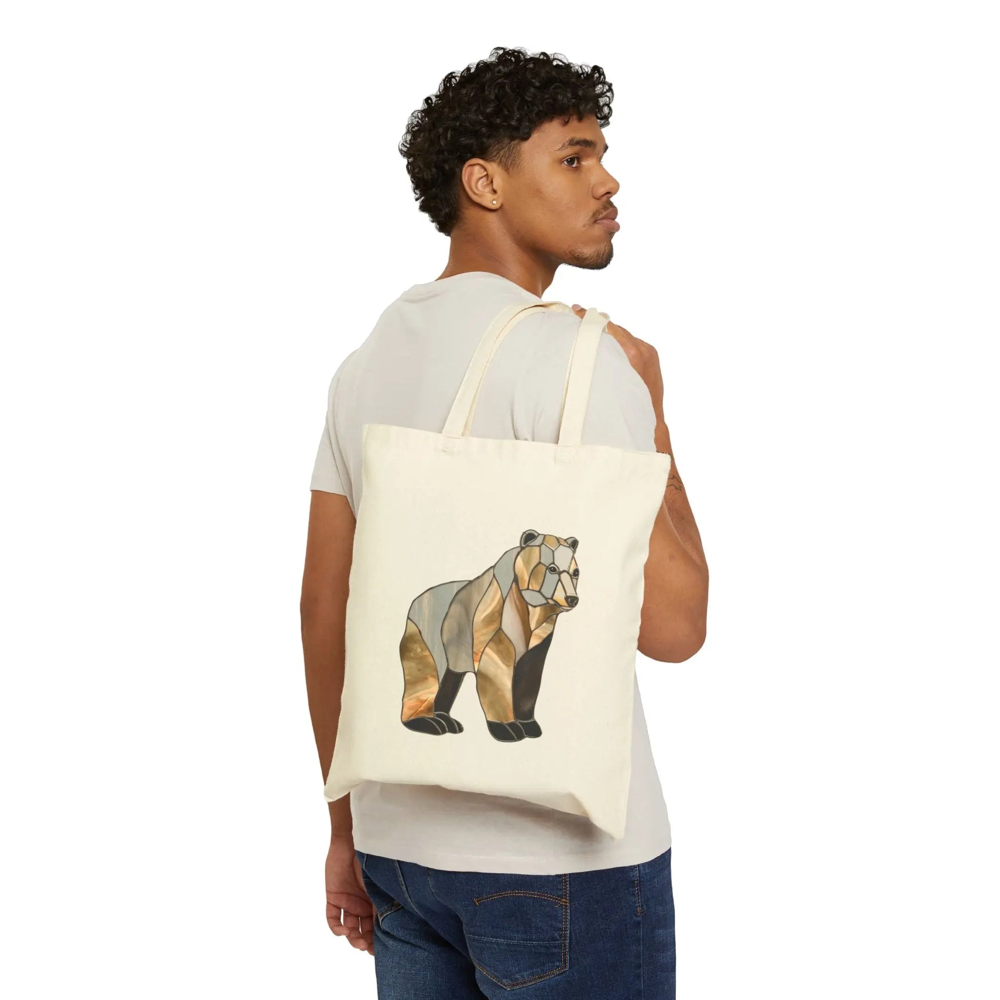 A person in a white t-shirt and jeans carries the Stained Glass Bear Cotton Canvas Tote Bag over their shoulder. The bag showcases an abstract bear design in earth tones, inspired by stained glass, against a plain white background.