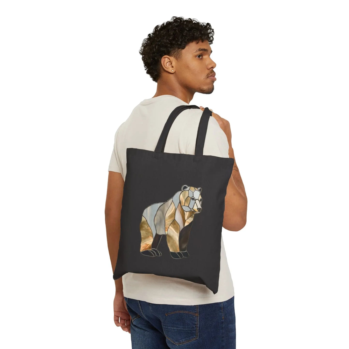 A man with curly hair in a beige t-shirt and jeans holds the Stained Glass Bear Cotton Canvas Tote Bag over his shoulder, showcasing its earthy-toned bear design.