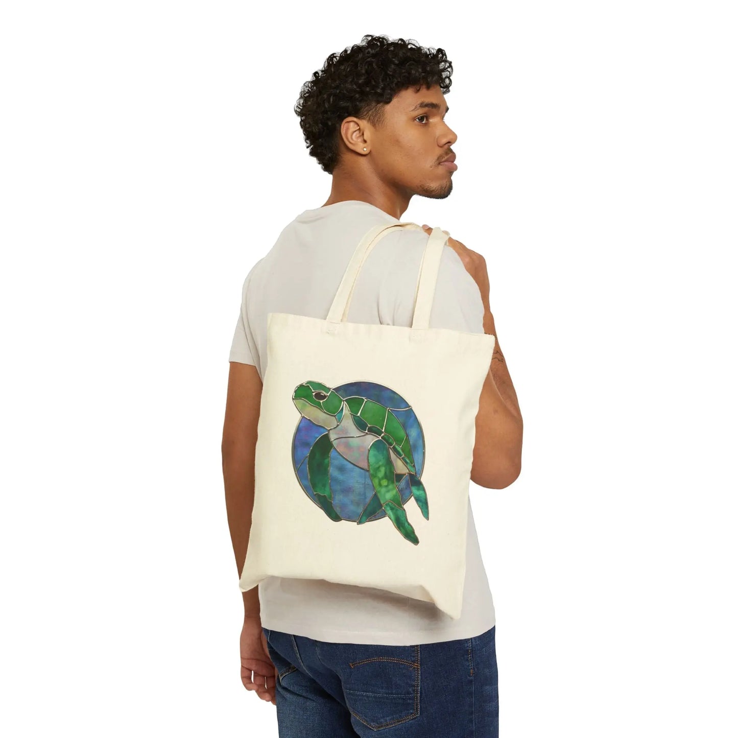A curly-haired person, dressed in a light beige t-shirt and blue jeans, holds the Stained Glass Turtle Cotton Canvas Tote Bag. It features a cream color with an illustration of a green turtle on a blue circular background.