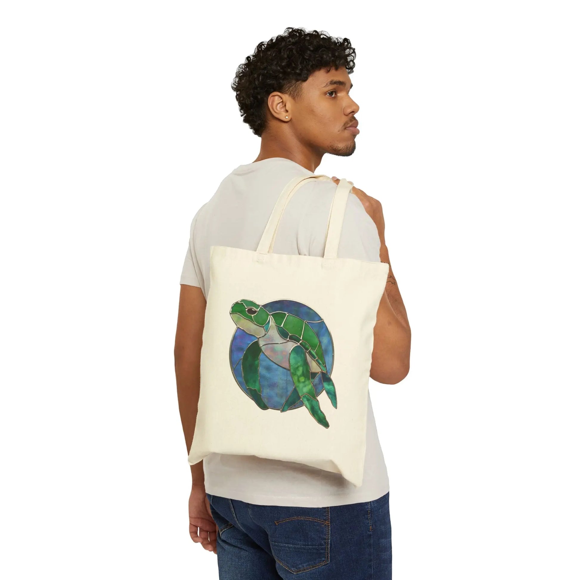 A curly-haired person, dressed in a light beige t-shirt and blue jeans, holds the Stained Glass Turtle Cotton Canvas Tote Bag. It features a cream color with an illustration of a green turtle on a blue circular background.