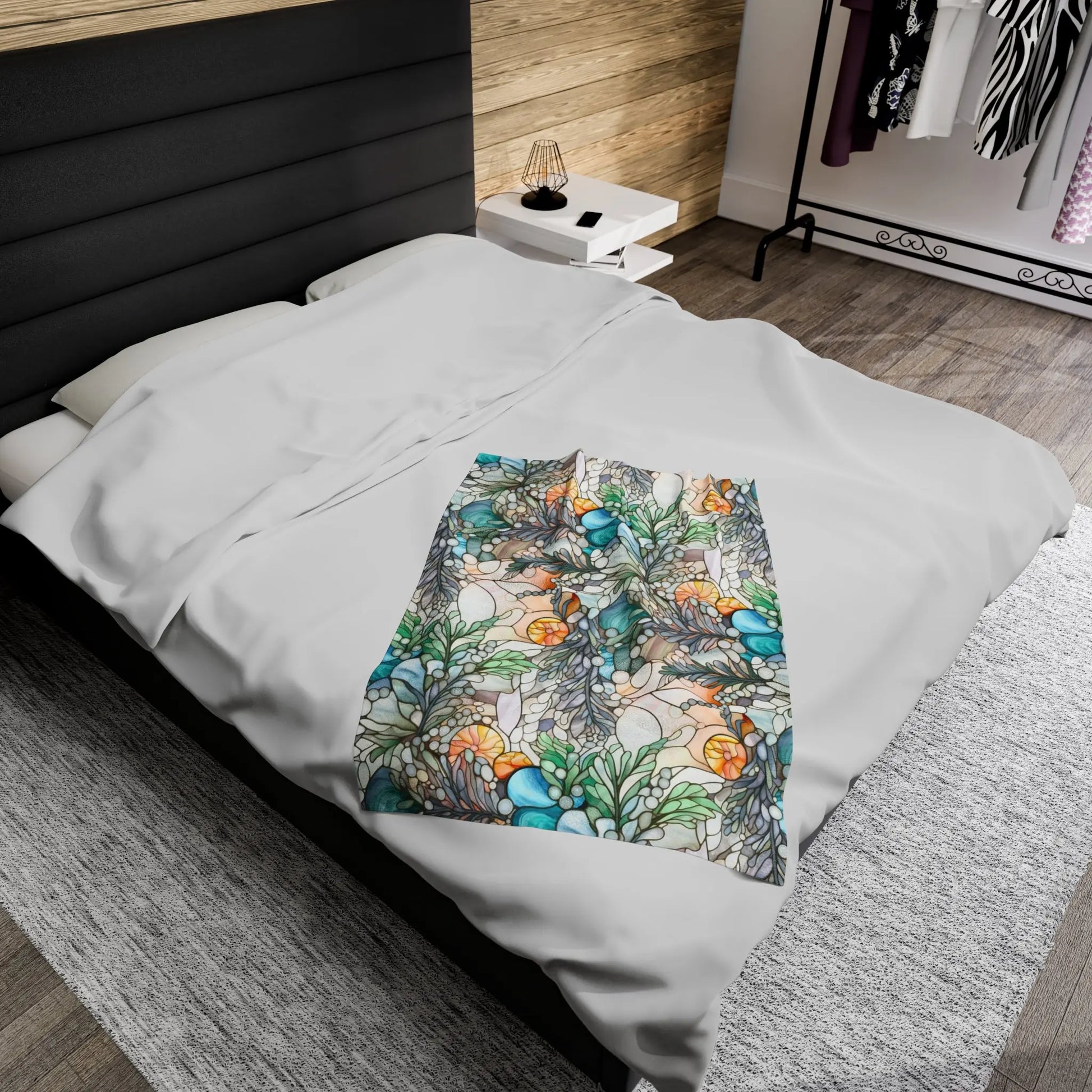 A neatly made bed with gray bedding features a Stained Glass Ferns Velveteen Soft Plush Blanket, showcasing corals and seashells. The room includes a wooden accent wall, bedside table with a lamp, clothing rack, and a gray rug on the wooden floor.