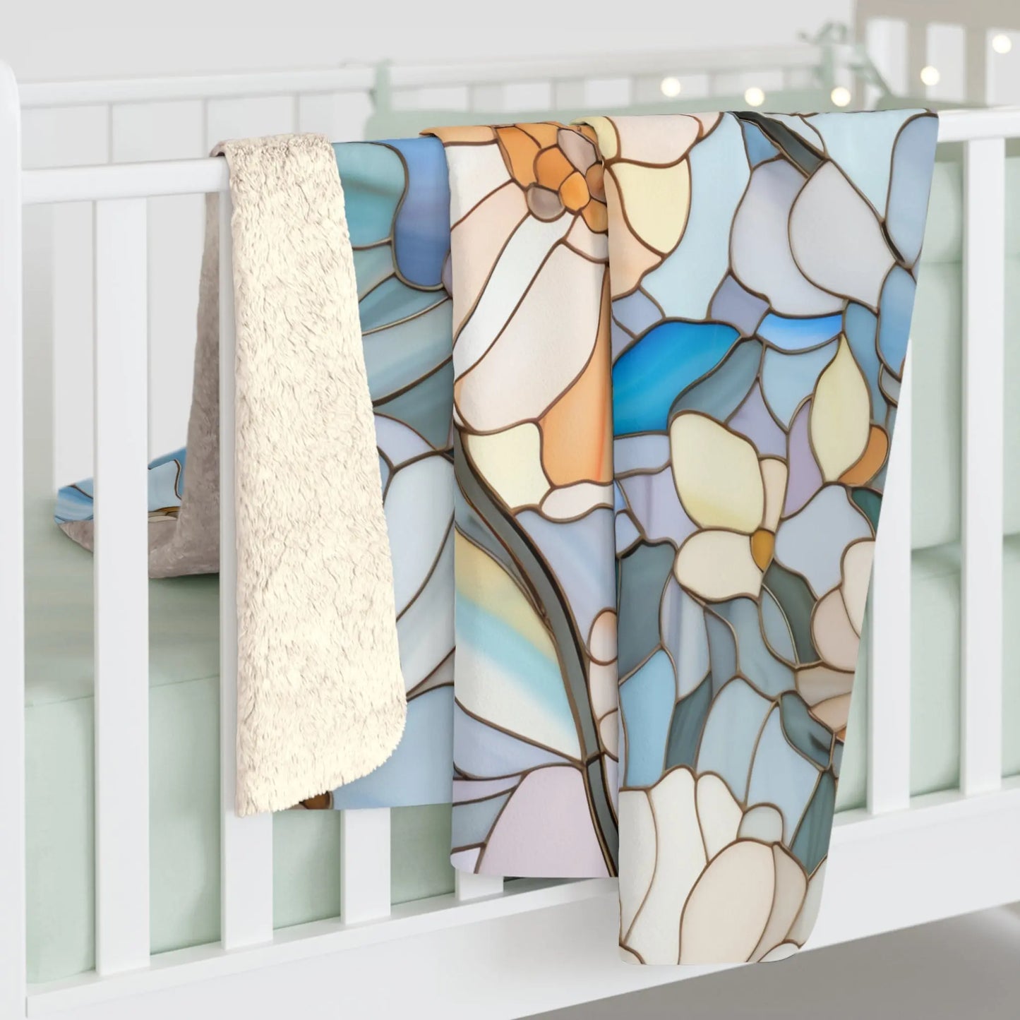 A baby crib with a light green mattress has two blankets draped over the side: one plush cream velveteen and a boho Stained Glass Flowers Sherpa Fleece Blanket, featuring a floral pattern in blue, orange, and beige.