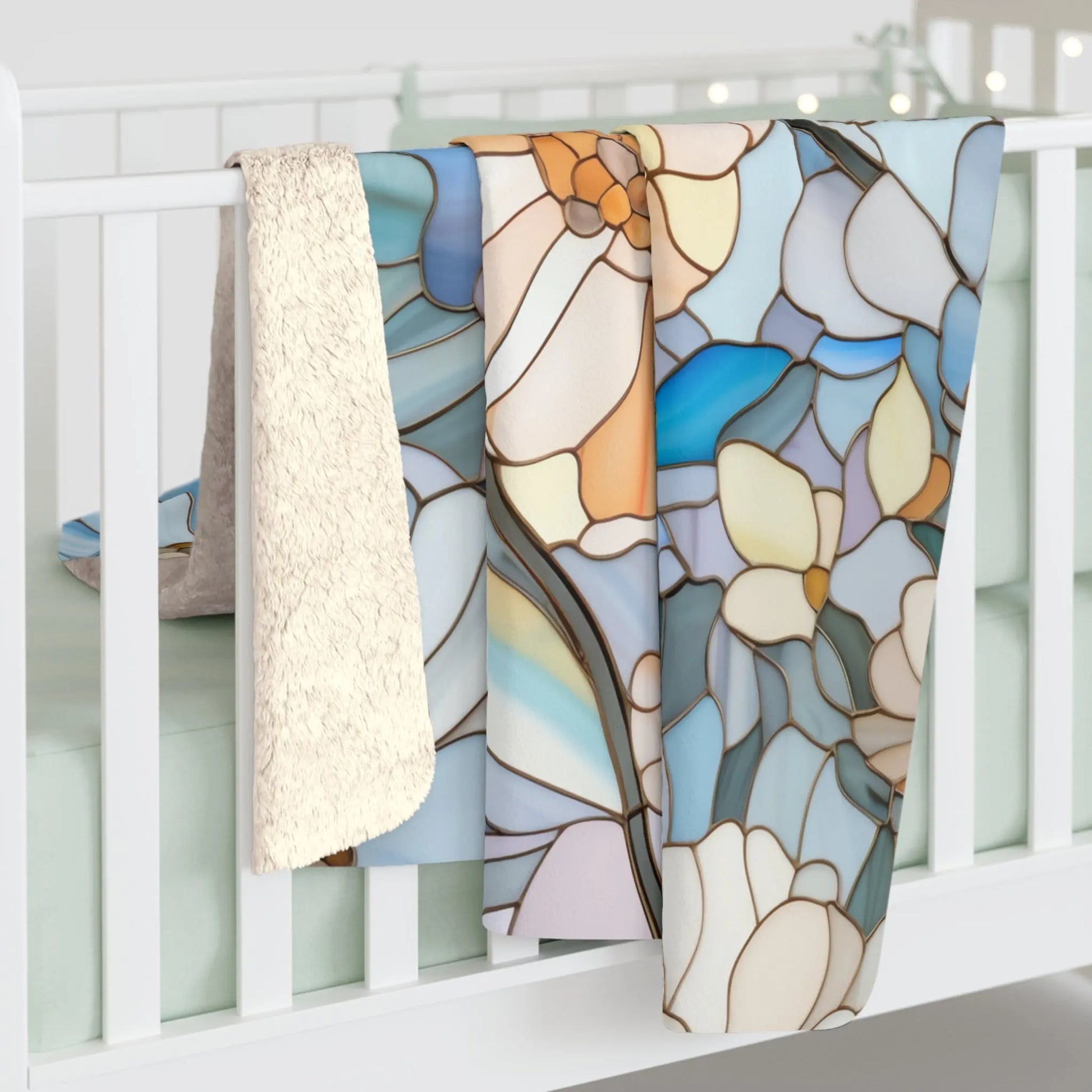 A baby crib with a light green mattress has two blankets draped over the side: one plush cream velveteen and a boho Stained Glass Flowers Sherpa Fleece Blanket, featuring a floral pattern in blue, orange, and beige.