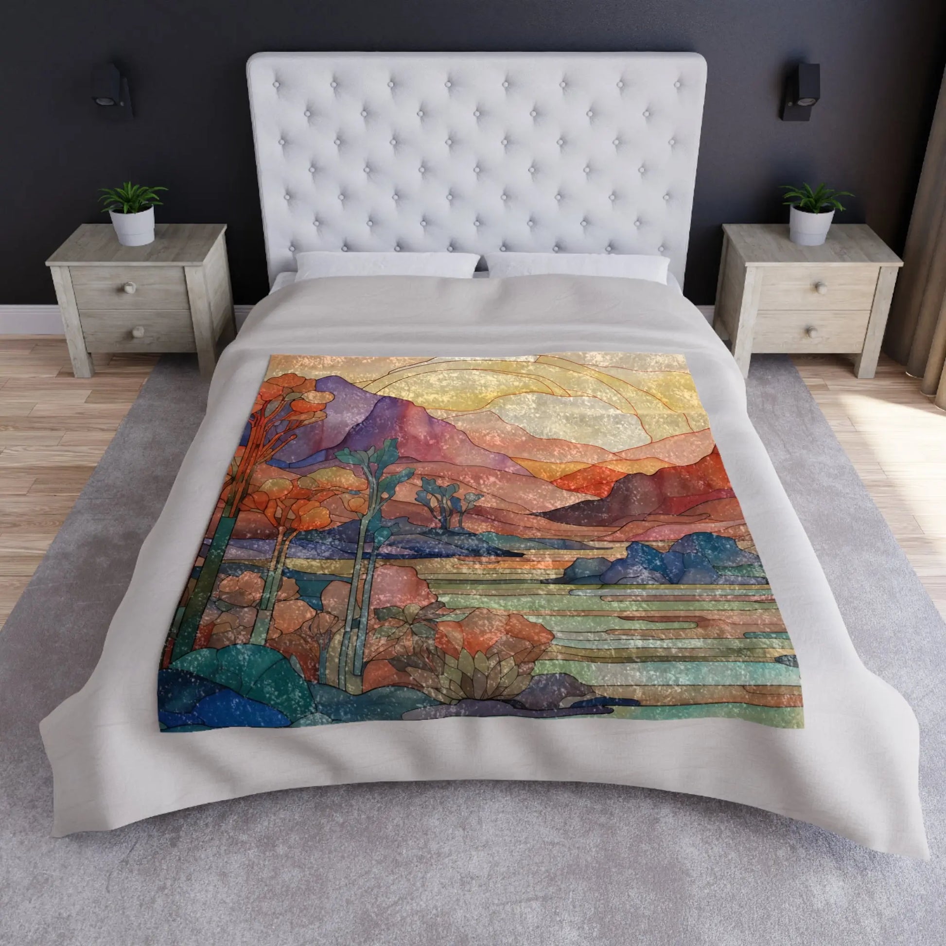 A bed adorned with a Stained Glass Desert Sunset Crushed Velvet Blanket (50x60) depicts mountains, trees, and a sunlit sky. The room has a grey carpet, two wooden bedside tables with potted plants, and dark walls.