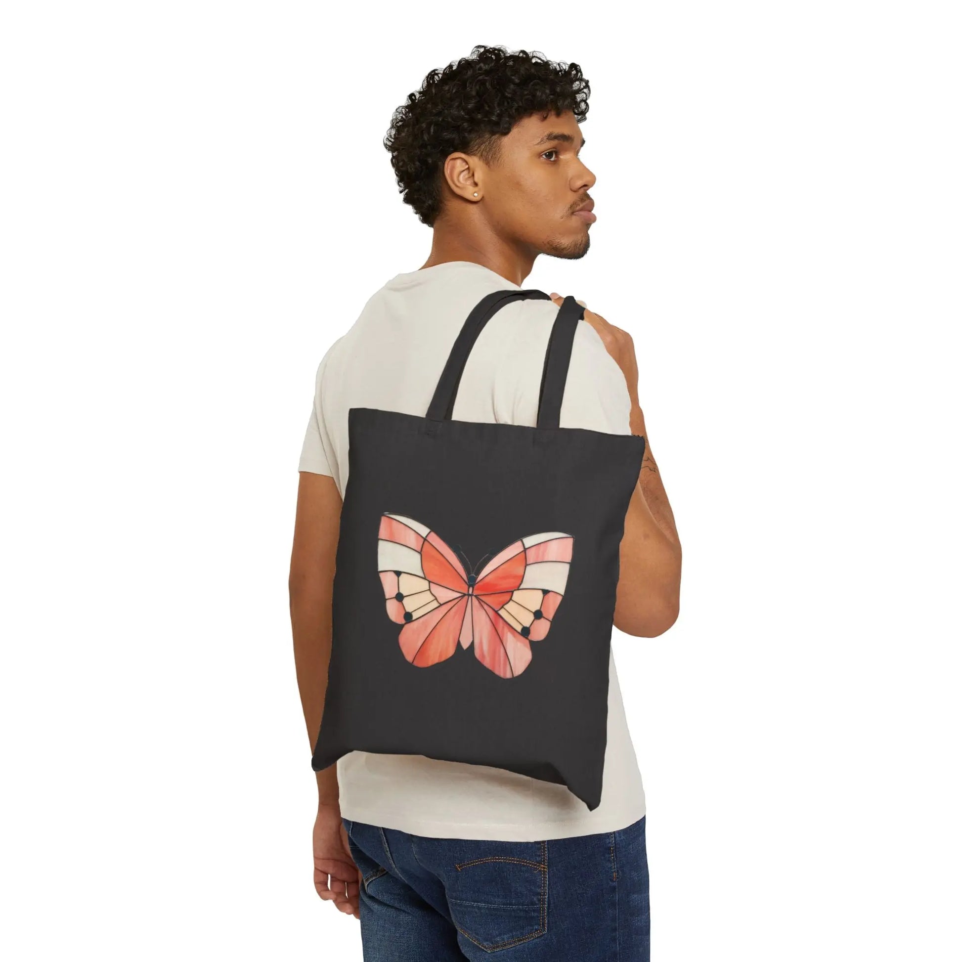 A person in a white shirt and jeans casually holds the Stained Glass Spring Butterfly Cotton Canvas Tote Bag over their shoulder. It showcases original artwork of a large, colorful butterfly in vibrant pink, orange, and black shades on a plain white background.