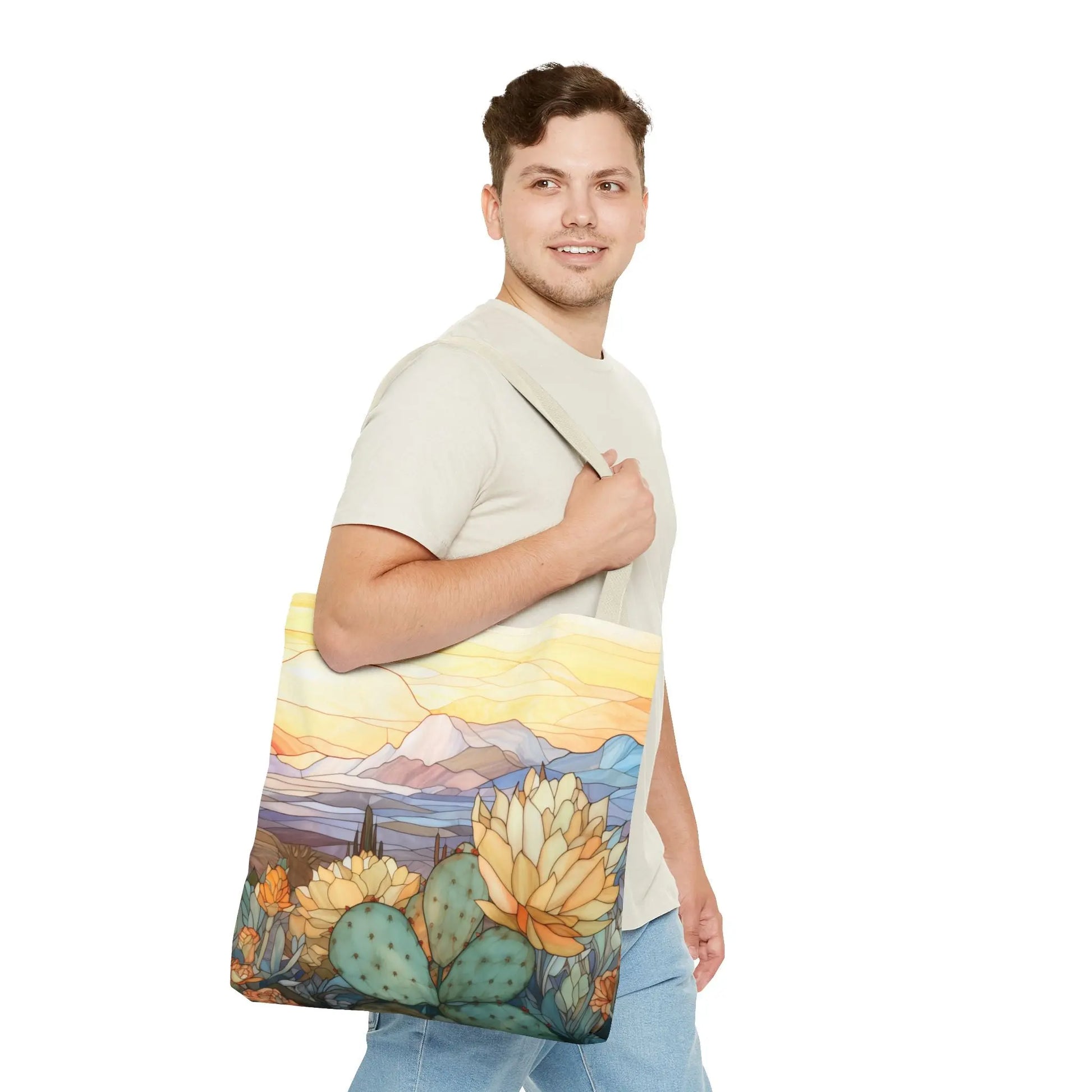 A man in a light beige t-shirt and jeans carries a colorful Stained Glass Desert Cactus Tote Bag, featuring an original artist design of cacti and mountains under a yellow and orange sky. Made from durable polyester, it resembles stained glass art. He stands against a plain white background.