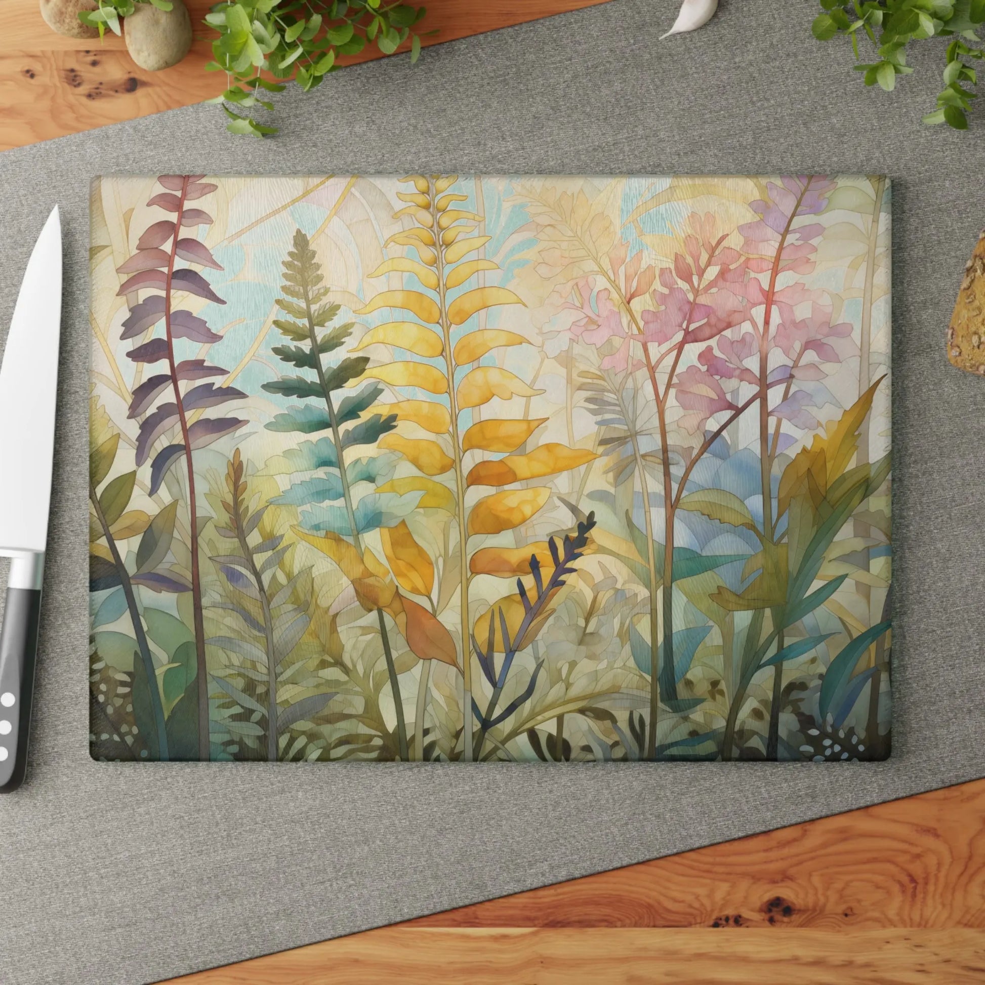 The Stained Glass Pastel Ferns Glass Cutting Board showcases a botanical design with pastel ferns, set against a grey fabric mat beside a knife and green potted plants on wood.