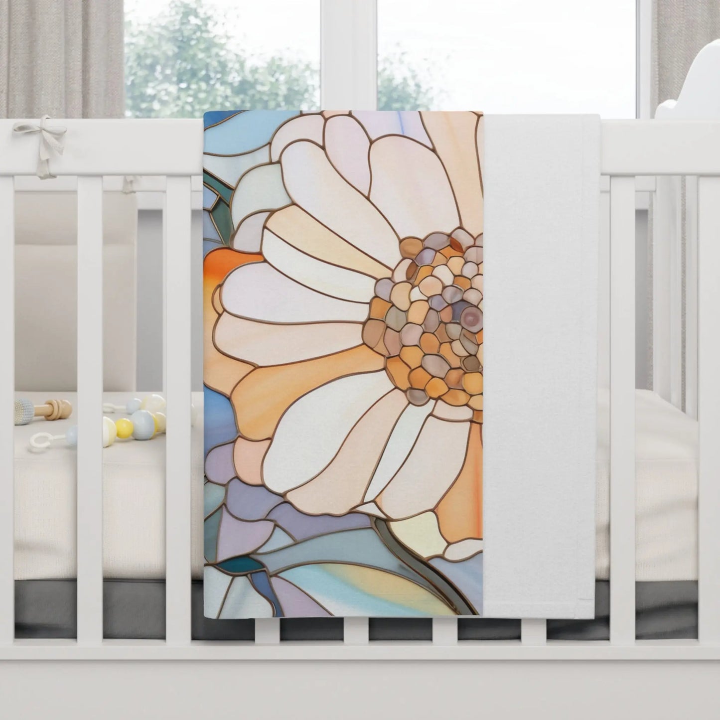 A white crib with a Stained Glass Flowers Soft Fleece Baby Blanket draped over the side exudes boho elegance. The blanket showcases an abstract floral design in orange, white, and blue hues, while pastel-colored baby rattles lie inside the crib.