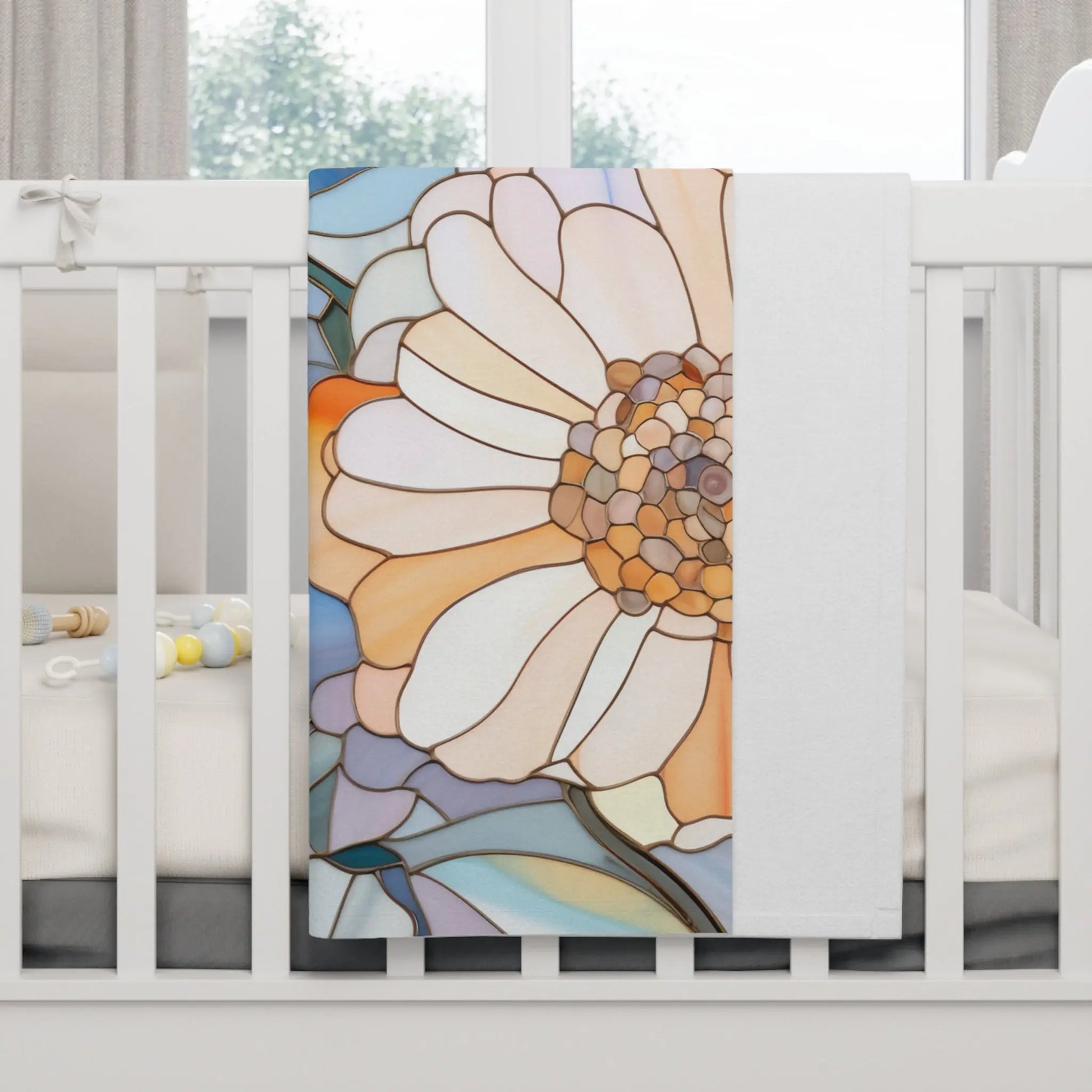 A white crib with a Stained Glass Flowers Soft Fleece Baby Blanket draped over the side exudes boho elegance. The blanket showcases an abstract floral design in orange, white, and blue hues, while pastel-colored baby rattles lie inside the crib.