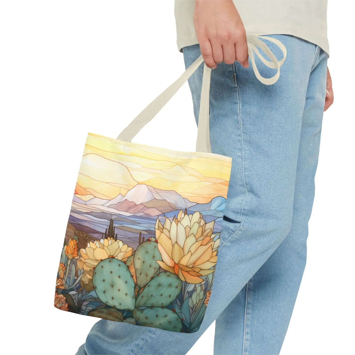 Someone in blue jeans carries the Stained Glass Desert Cactus Tote Bag, featuring an artist design of a vibrant desert scene. Colorful cacti, yellow flowers, and mountains under a golden sky stand out against the durable polyester material.