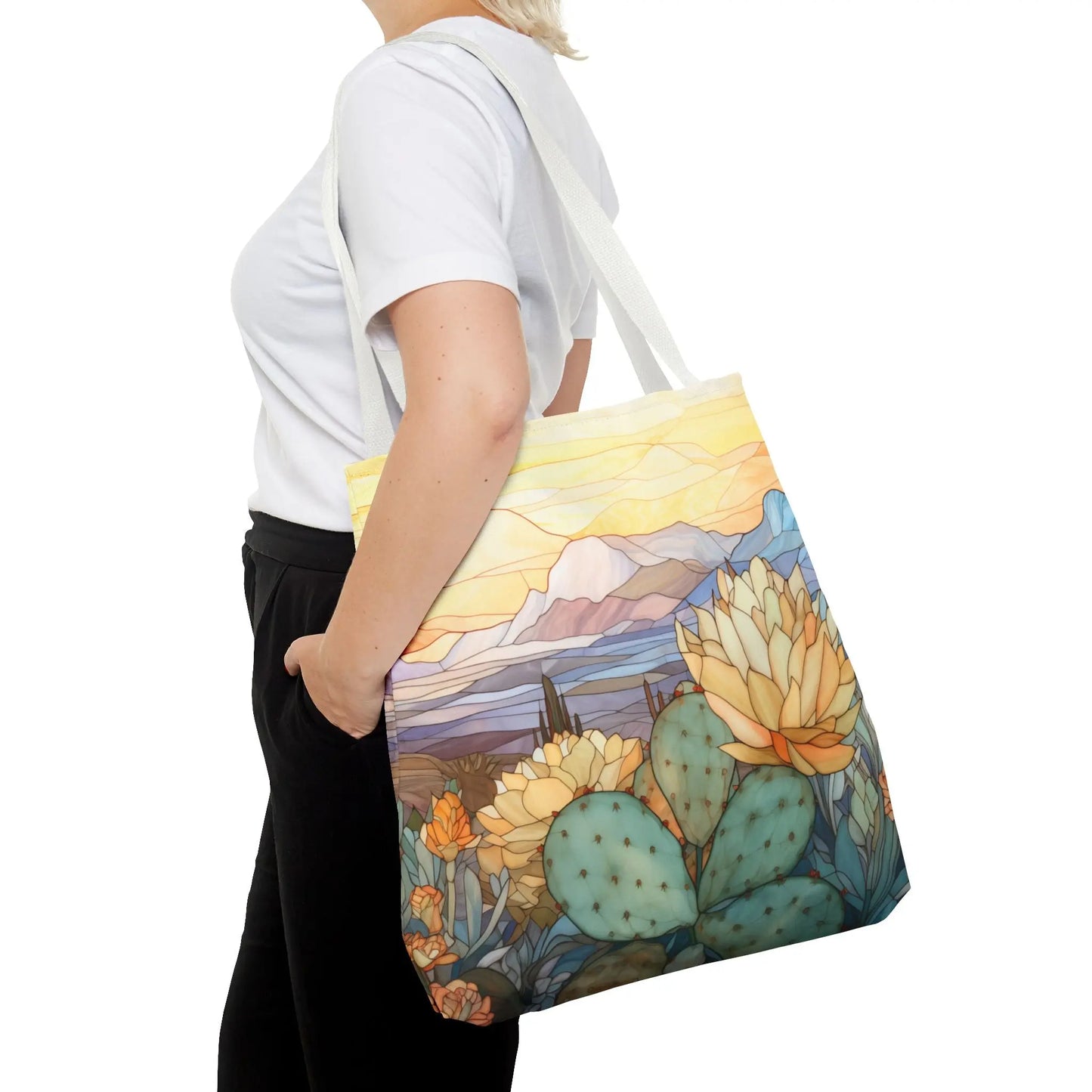 A person in a white shirt and black pants carries a vibrant Stained Glass Desert Cactus Tote Bag showcasing an original desert landscape design with cacti, flowers, mountains, and a sunset sky, all crafted from durable polyester.