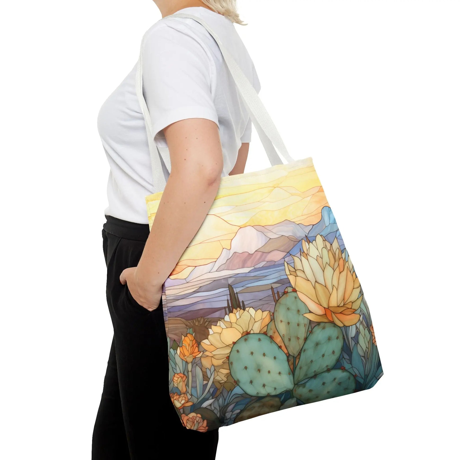 A person in a white shirt and black pants carries a vibrant Stained Glass Desert Cactus Tote Bag showcasing an original desert landscape design with cacti, flowers, mountains, and a sunset sky, all crafted from durable polyester.
