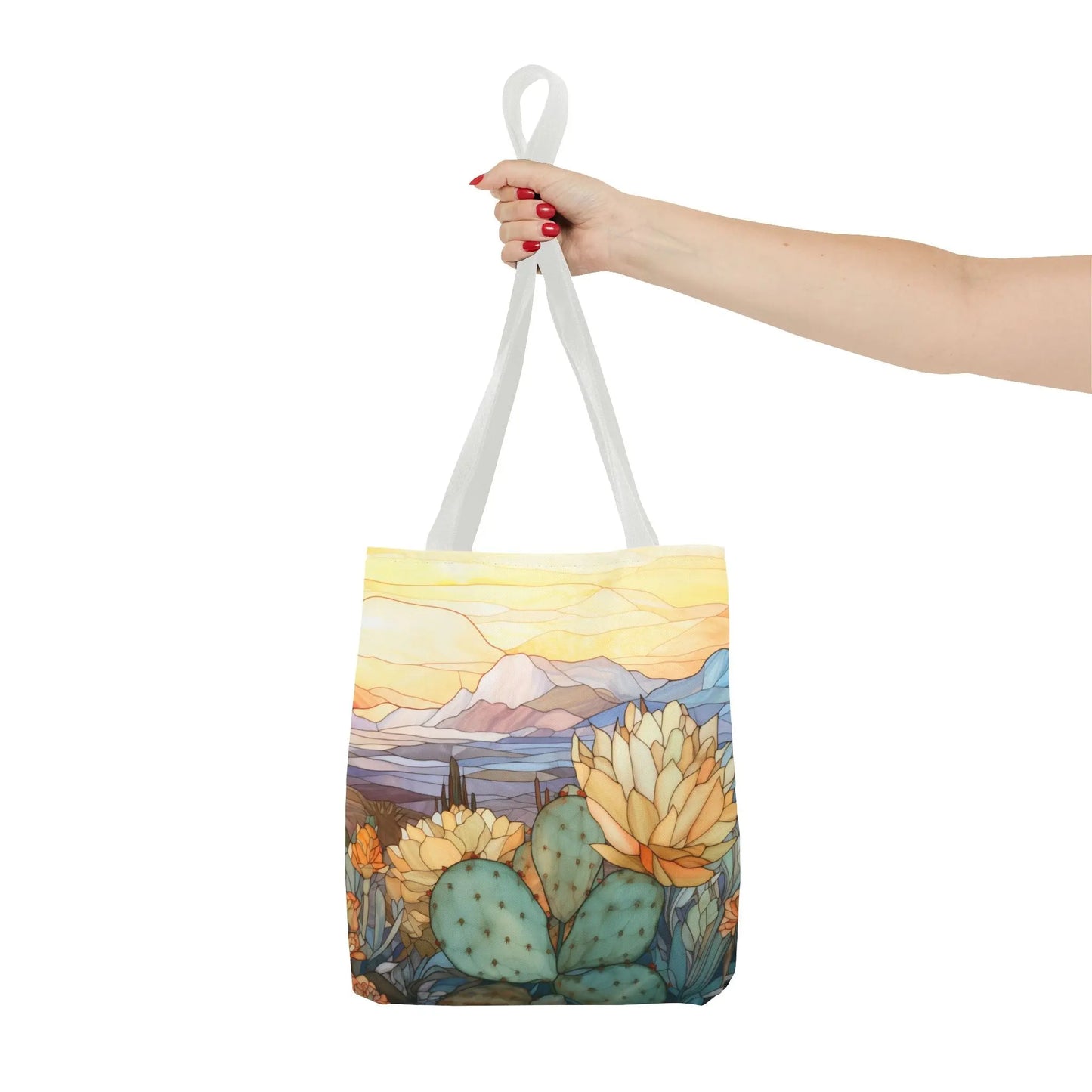 A hand with red nail polish showcases the Stained Glass Desert Cactus Tote Bag, featuring an original design of a desert scene with blooming cacti and sunset-lit mountains. Made from durable polyester, it has white straps and a vibrant, colorful look. Available in 3 sizes.
