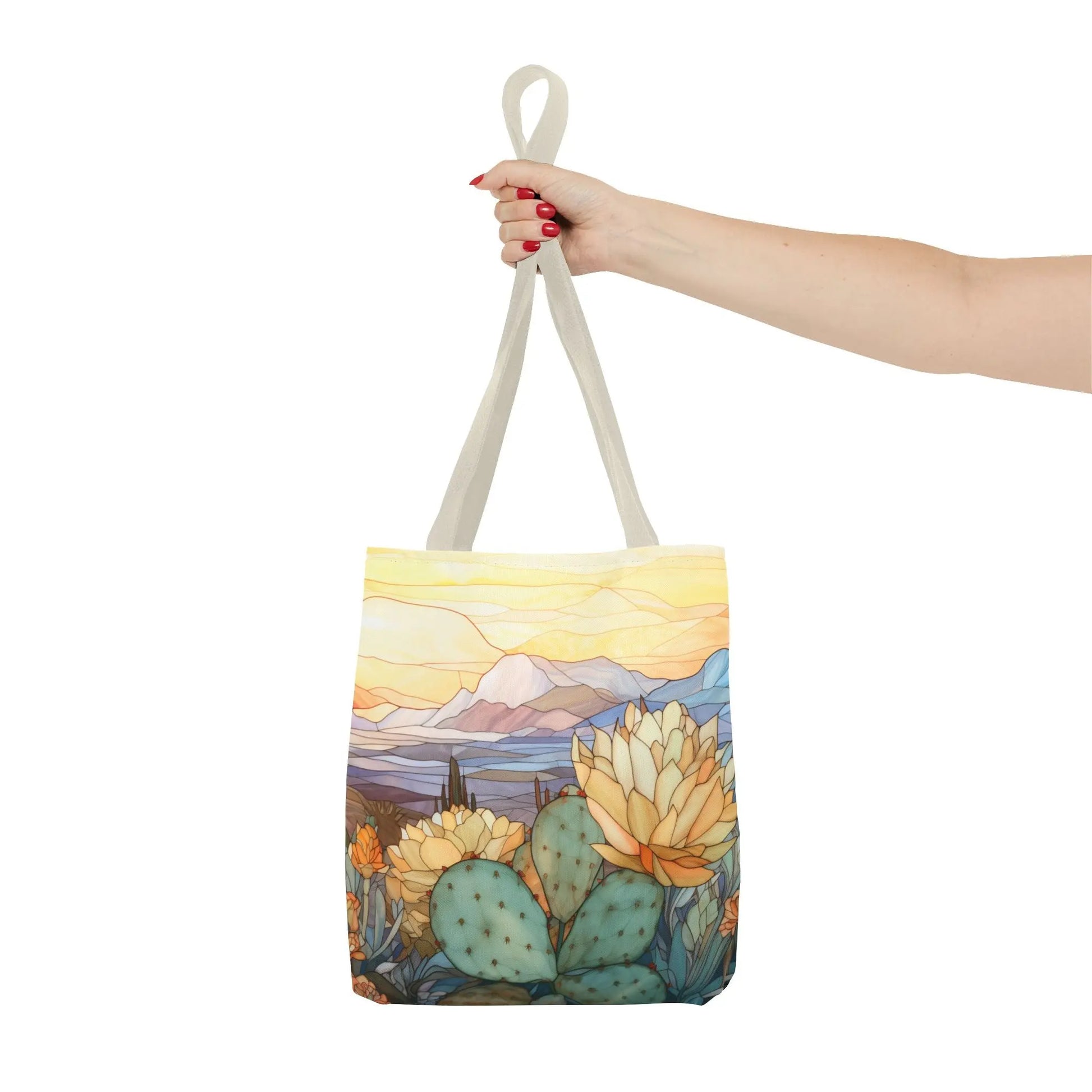 A hand with red nail polish holds the Stained Glass Desert Cactus Tote Bag, showcasing a desert landscape with cacti, blooming flowers, mountains, and a sunset sky. Made from durable polyester, this accessory is as stylish as it is resilient.