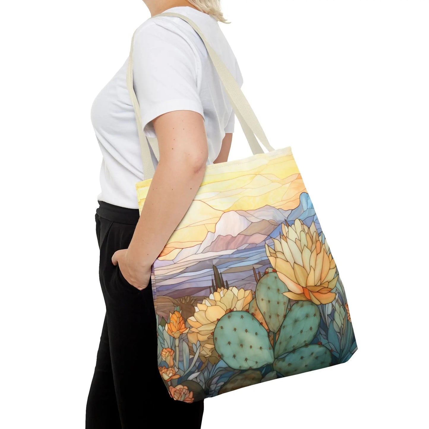 Someone wears a white t-shirt and black pants while carrying the Stained Glass Desert Cactus Tote Bag, made of durable polyester. The bag features an original design with cacti, flowers, and mountains under a sunset sky.