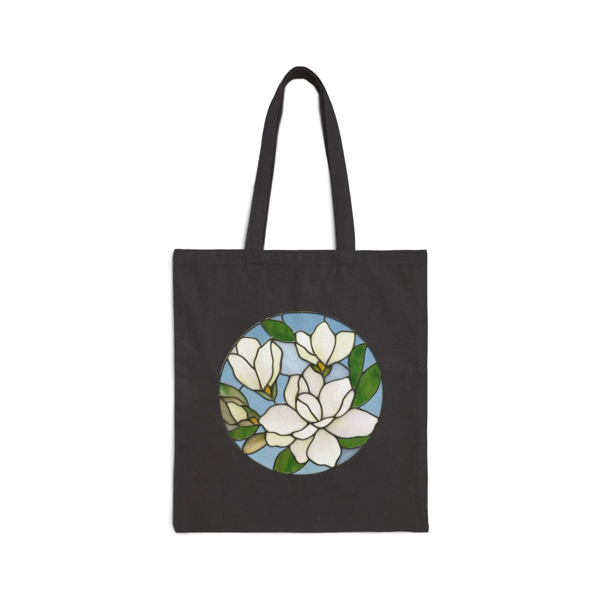The Stained Glass Magnolia Cotton Canvas Tote Bag by Noelle Barnes features a black bag with a central stained glass design of white magnolias and green leaves on a light blue background. It has two long handles for easy carrying.