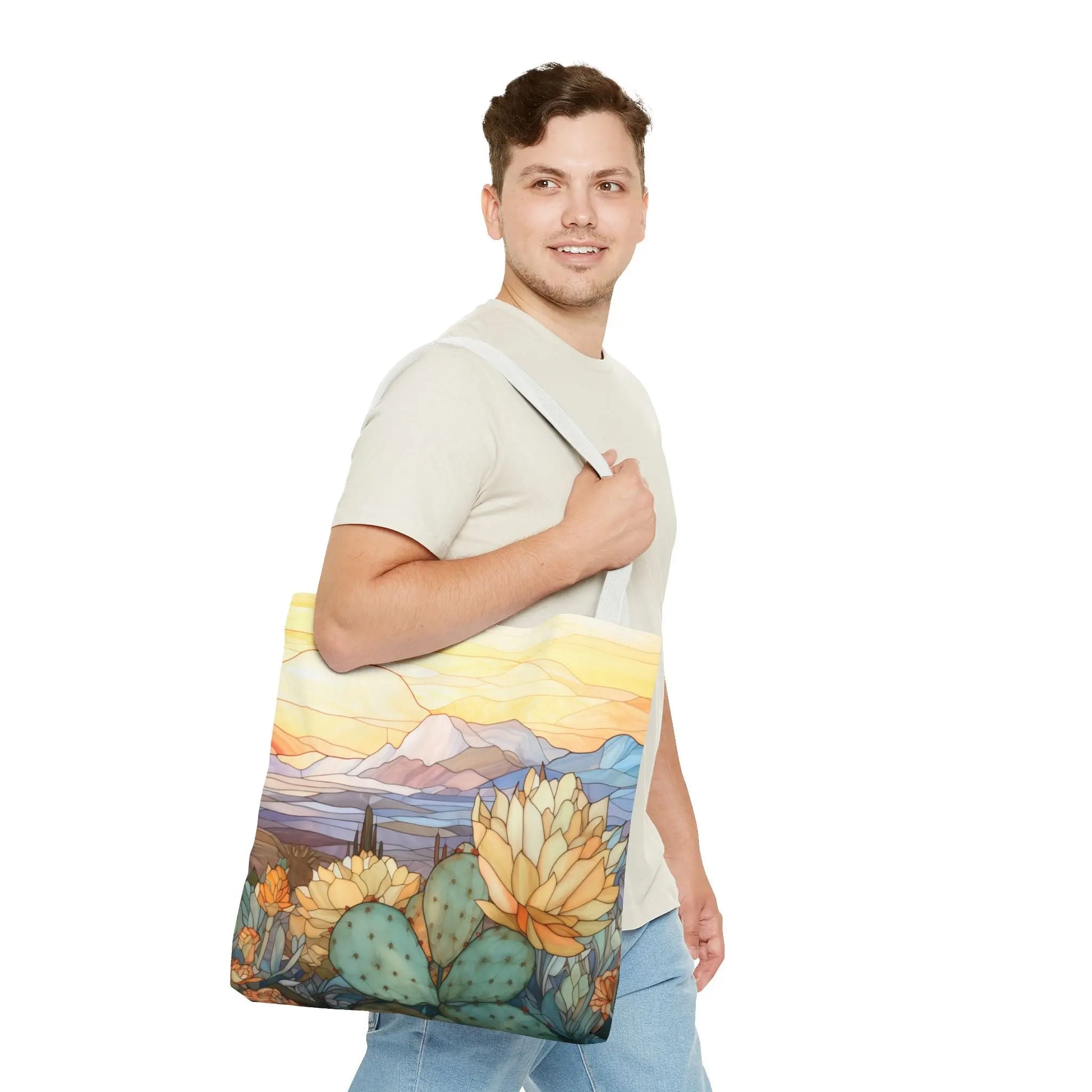 A person in a light beige t-shirt and blue jeans carries a large Stained Glass Desert Cactus Tote Bag, showcasing an original artists colorful desert landscape with mountains, cacti, and soft sky hues. Made of durable polyester, it stands out against the plain white background.
.