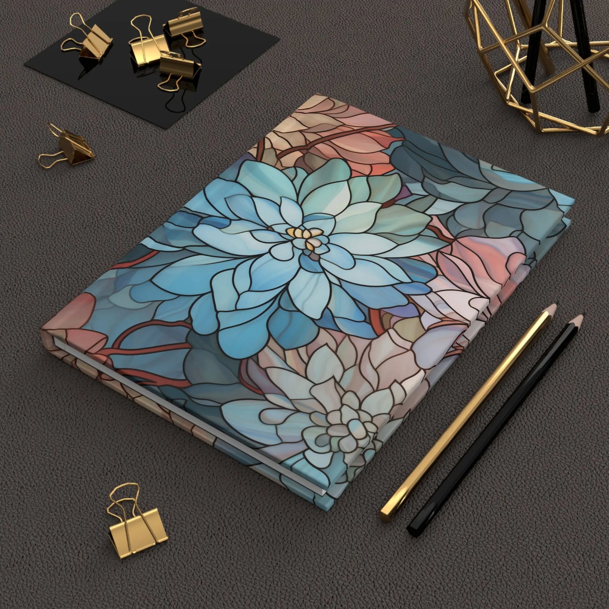 A Stained Glass Dark Flowers Hardcover Journal rests stylishly on a textured surface, surrounded by gold and black pencils, a geometric stained-glass-like decoration, and gold binder clips atop a black sheet.