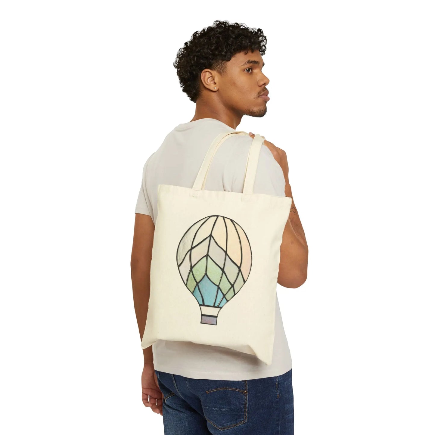 A person with curly hair turns slightly, holding a beige Stained Glass Hot Air Balloon Cotton Canvas Tote Bag. They wear a light gray shirt and jeans, set against a plain white background.