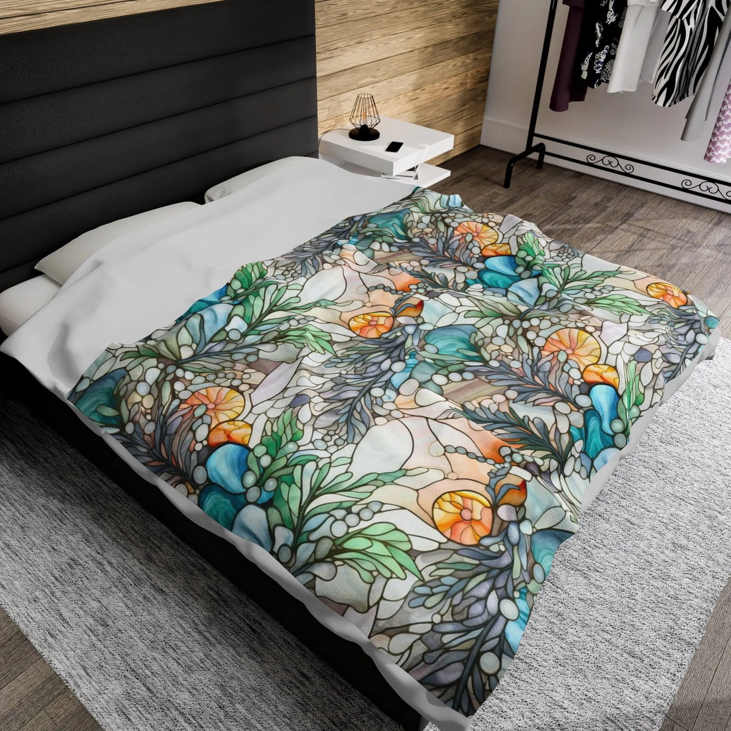 A bed adorned with a Stained Glass Ferns Velveteen Soft Plush Blanket, showcasing vibrant floral and leaf patterns in green, orange, and blue. The room features wooden walls, a nightstand with a lamp, and a clothing rack in the background.