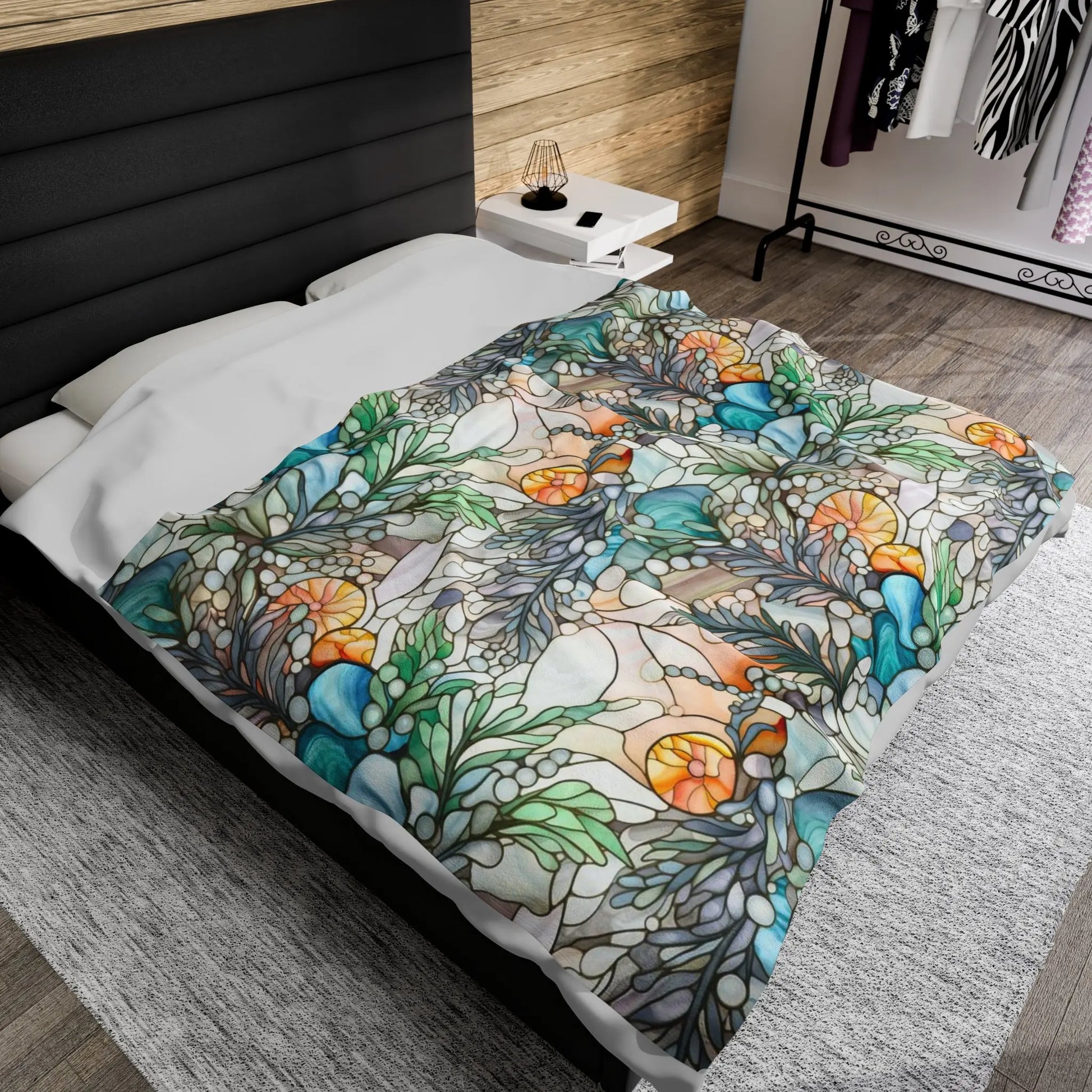 A bed adorned with a Stained Glass Ferns Velveteen Soft Plush Blanket, showcasing vibrant floral and leaf patterns in green, orange, and blue. The room features wooden walls, a nightstand with a lamp, and a clothing rack in the background.