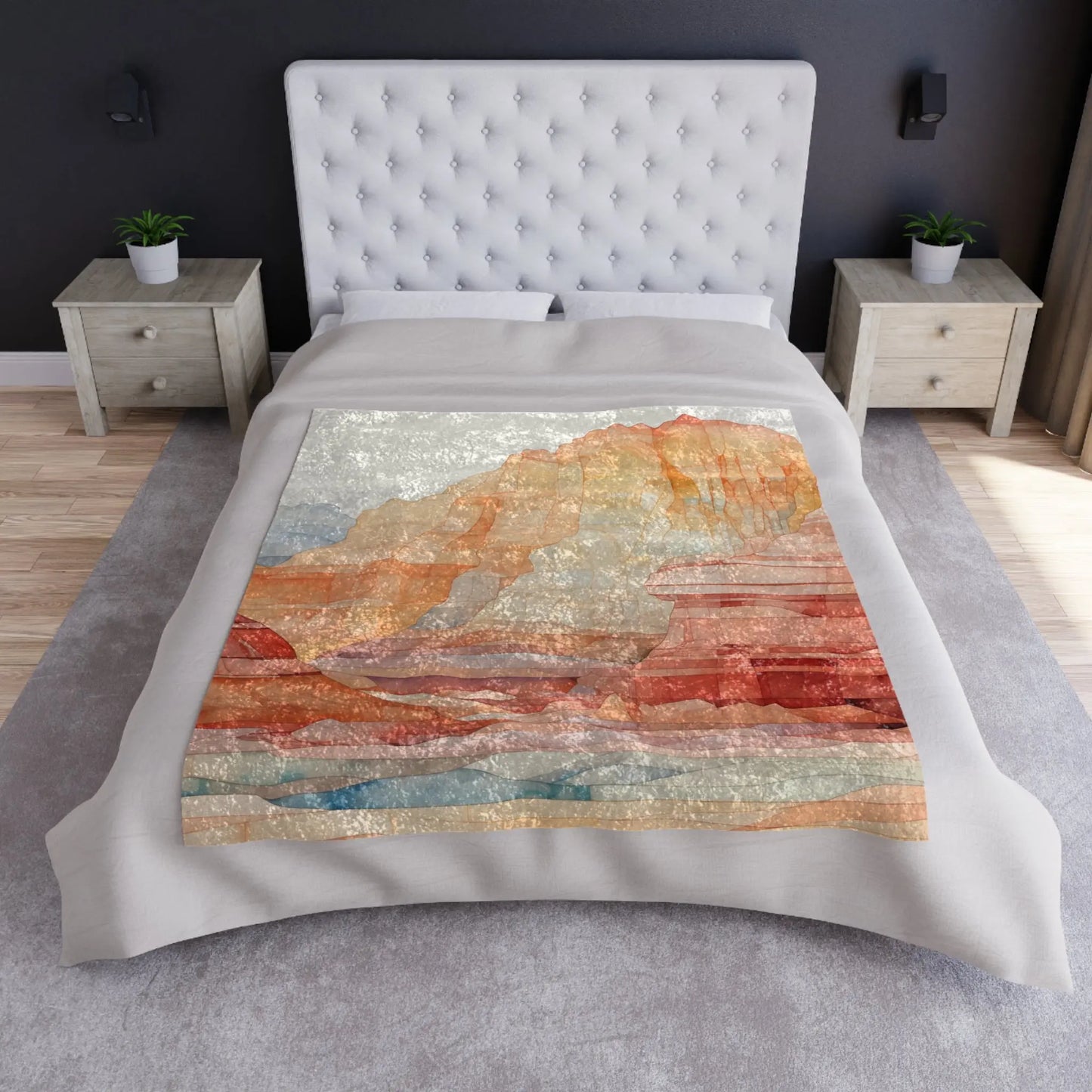 A bedroom featuring a tufted headboard and bed covered with a Stained Glass Sedona Red Rocks Crushed Velvet & Fleece Blanket – 50x60, displaying an abstract mountain landscape. Two bedside tables with small plants flank the bed, enhancing the neutral color palette.