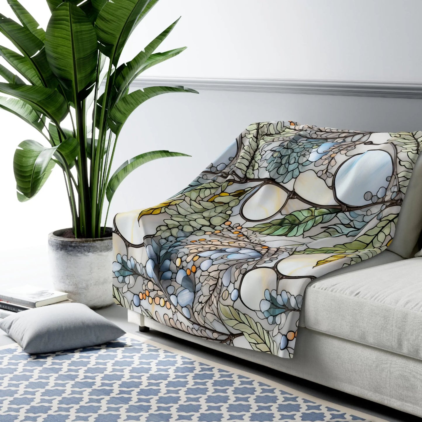 A cozy white sofa hosts a Stained Glass Nouveau Ferns Sherpa Fleece Blanket with leaf and abstract patterns. A tall green plant stands nearby, while a blue and white geometric rug complements two floor cushions.