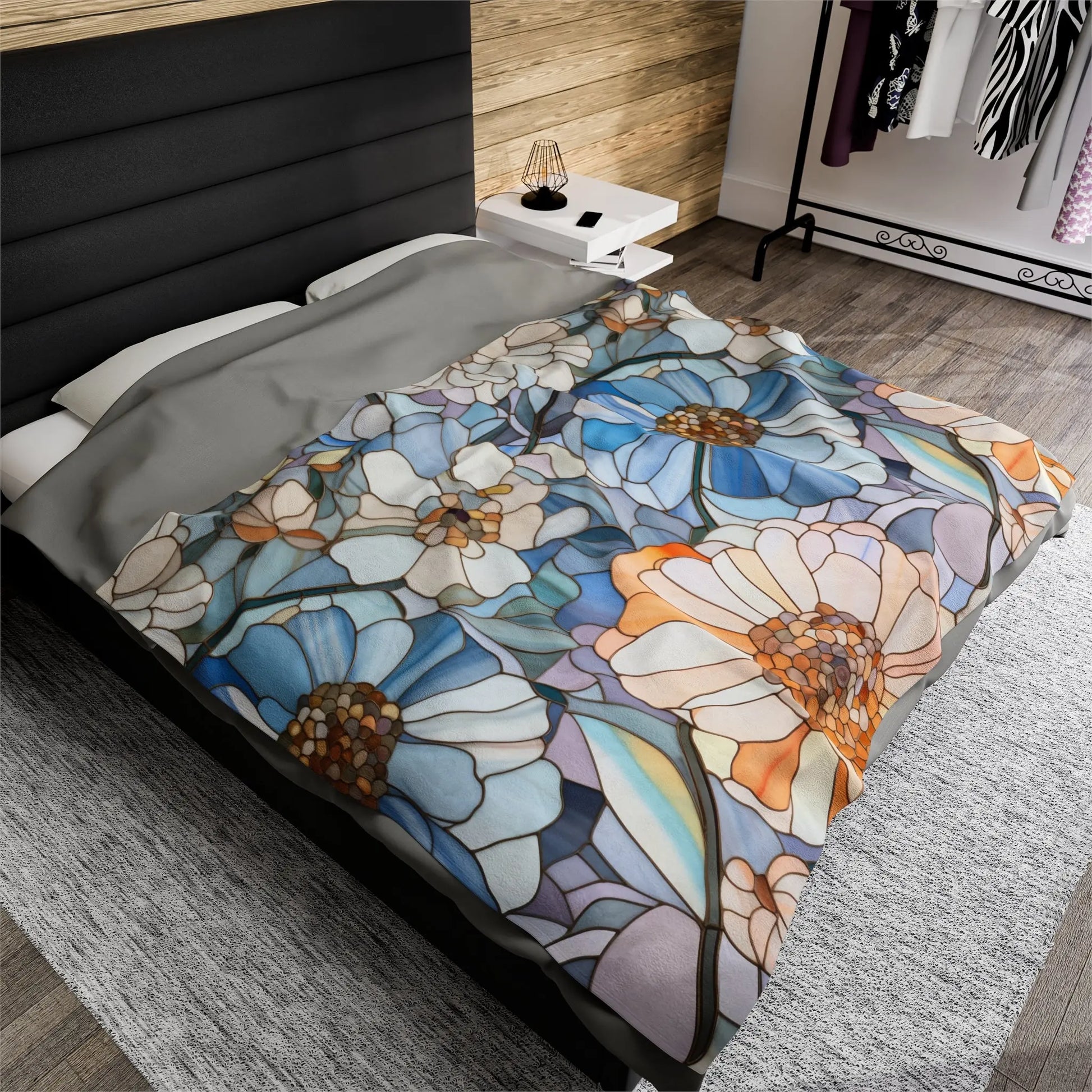 The bed is adorned with the Stained Glass Flowers Plush Blanket in blue, white, and orange. The room includes a wooden accent wall, a grey rug, a clothes-filled wardrobe, and a nightstand with a lamp next to the cozy arrangement.
