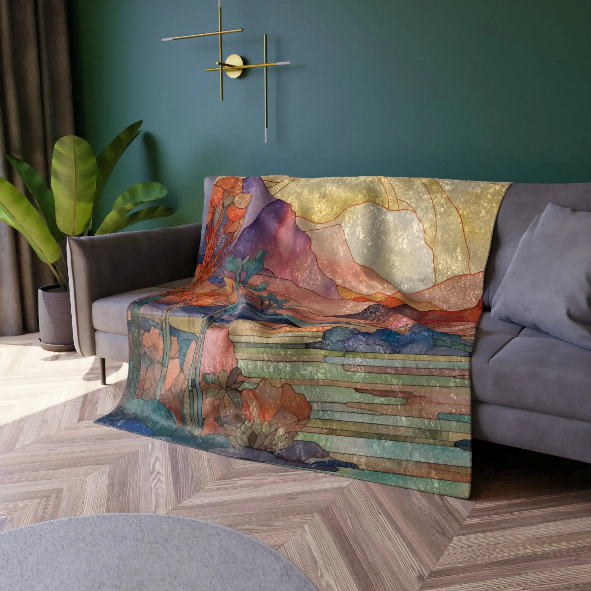 The Stained Glass Desert Sunset Crushed Velvet Blanket – 50x60 drapes over a gray couch in a room with green walls, a gold geometric wall decoration, a potted plant, and wooden flooring topped with a circular rug. Soft light warmly illuminates the space.