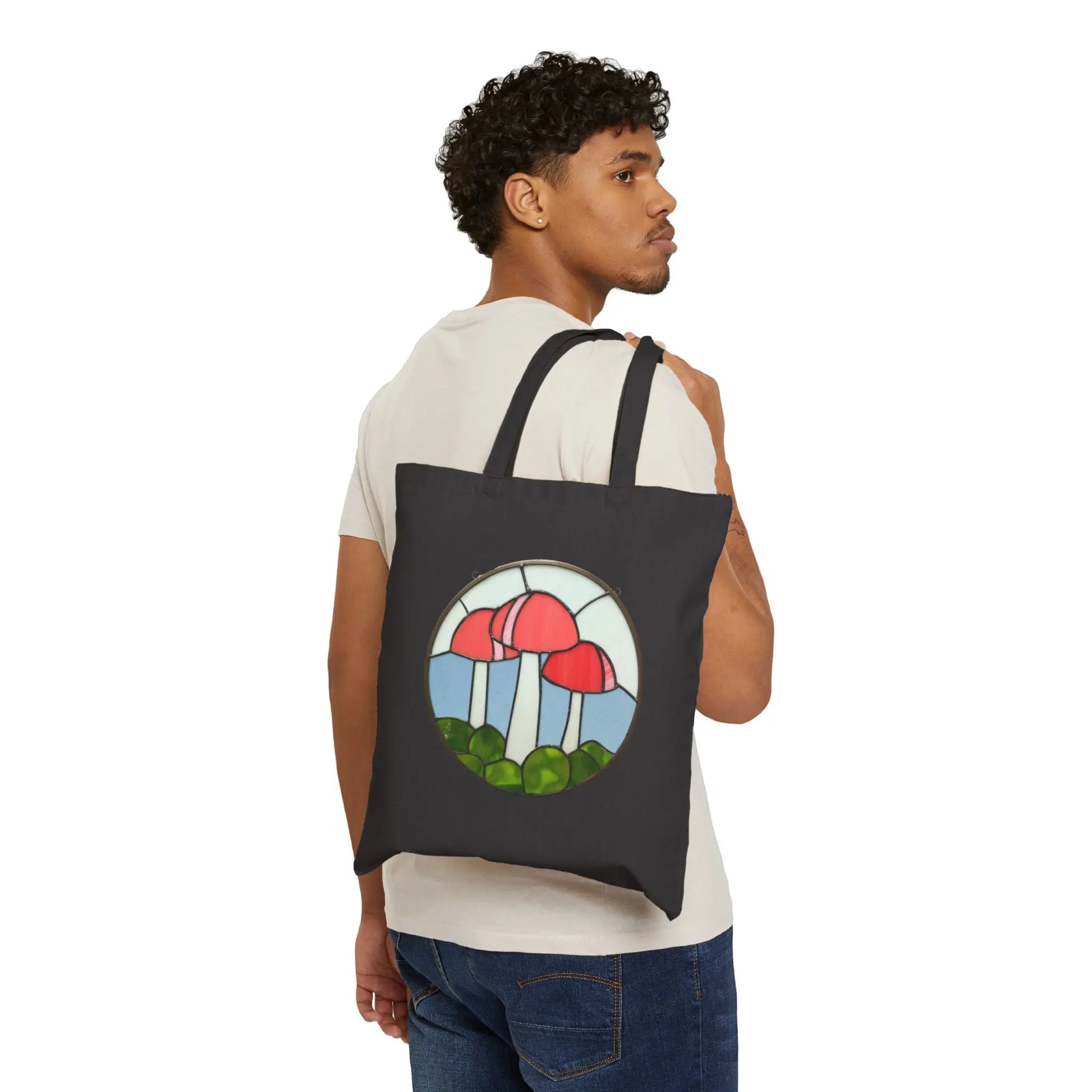 A person in a white shirt and jeans carries a black canvas tote bag featuring the Stained Glass Mushrooms design with red mushrooms and greenery.