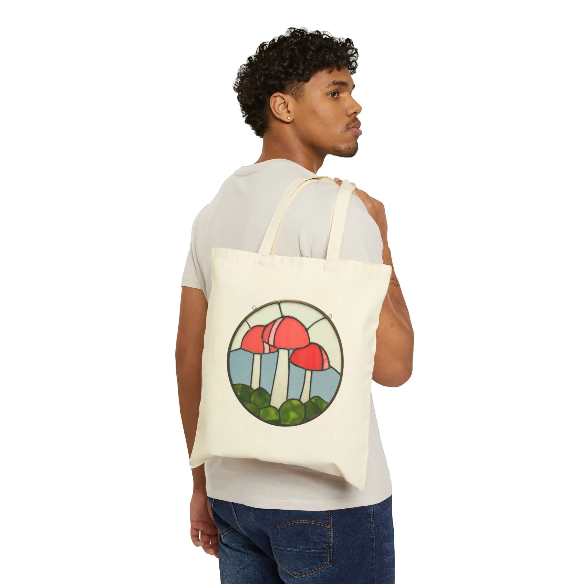 Noelle Barnes, with curly hair, wears a white t-shirt and jeans, effortlessly carrying over her shoulder the Stained Glass Mushrooms Cotton Canvas Tote Bag. It features a vibrant stained glass design with red mushrooms and lush green foliage.