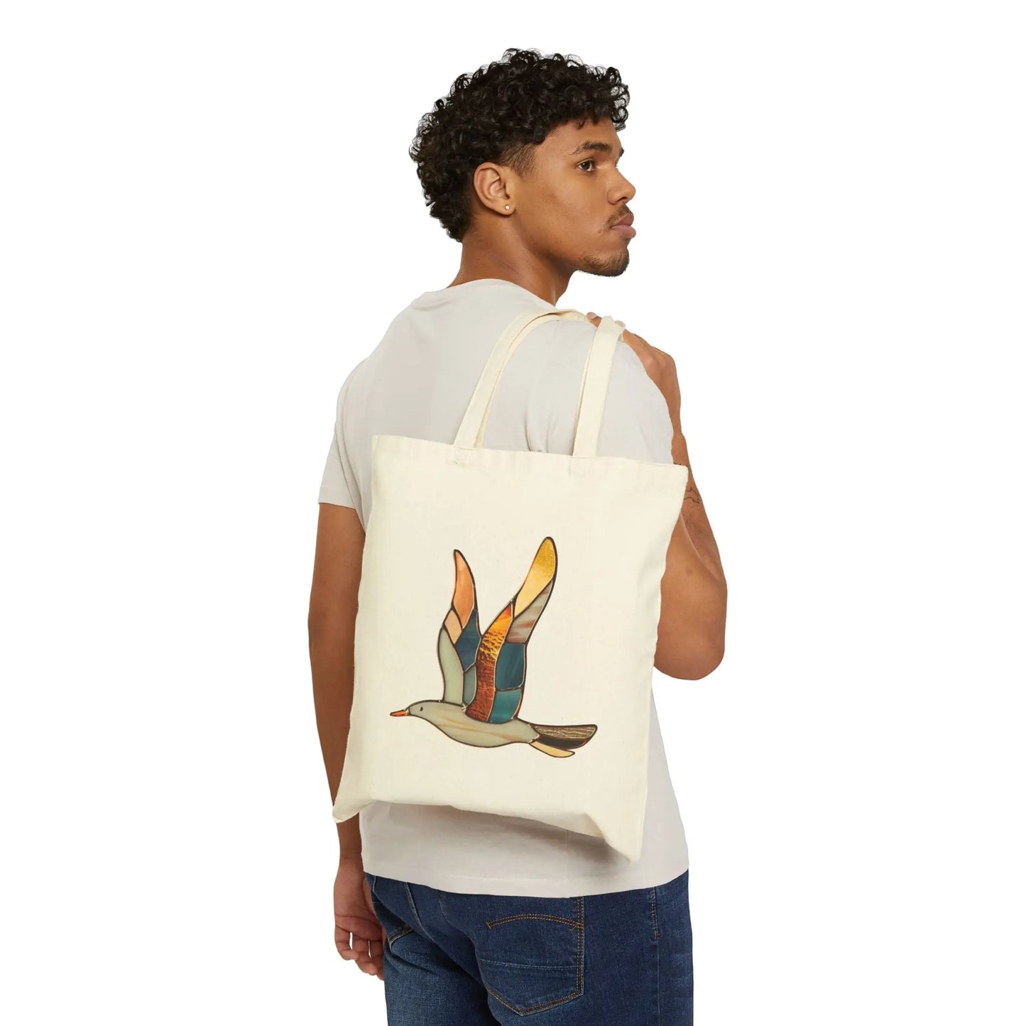 A curly-haired person in a white t-shirt and jeans carries the Stained Glass Seagull Cotton Canvas Tote Bag, featuring a colorful bird on a beige background, over their shoulder.