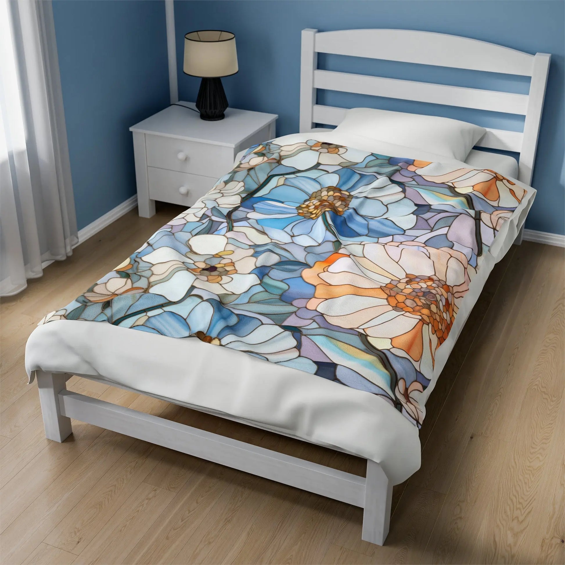 A white bed features the Stained Glass Flowers Plush Blanket with vibrant blue, orange, and white floral patterns. The room has a wooden floor, a lamp on a white bedside table, light blue walls, and a window to the left.