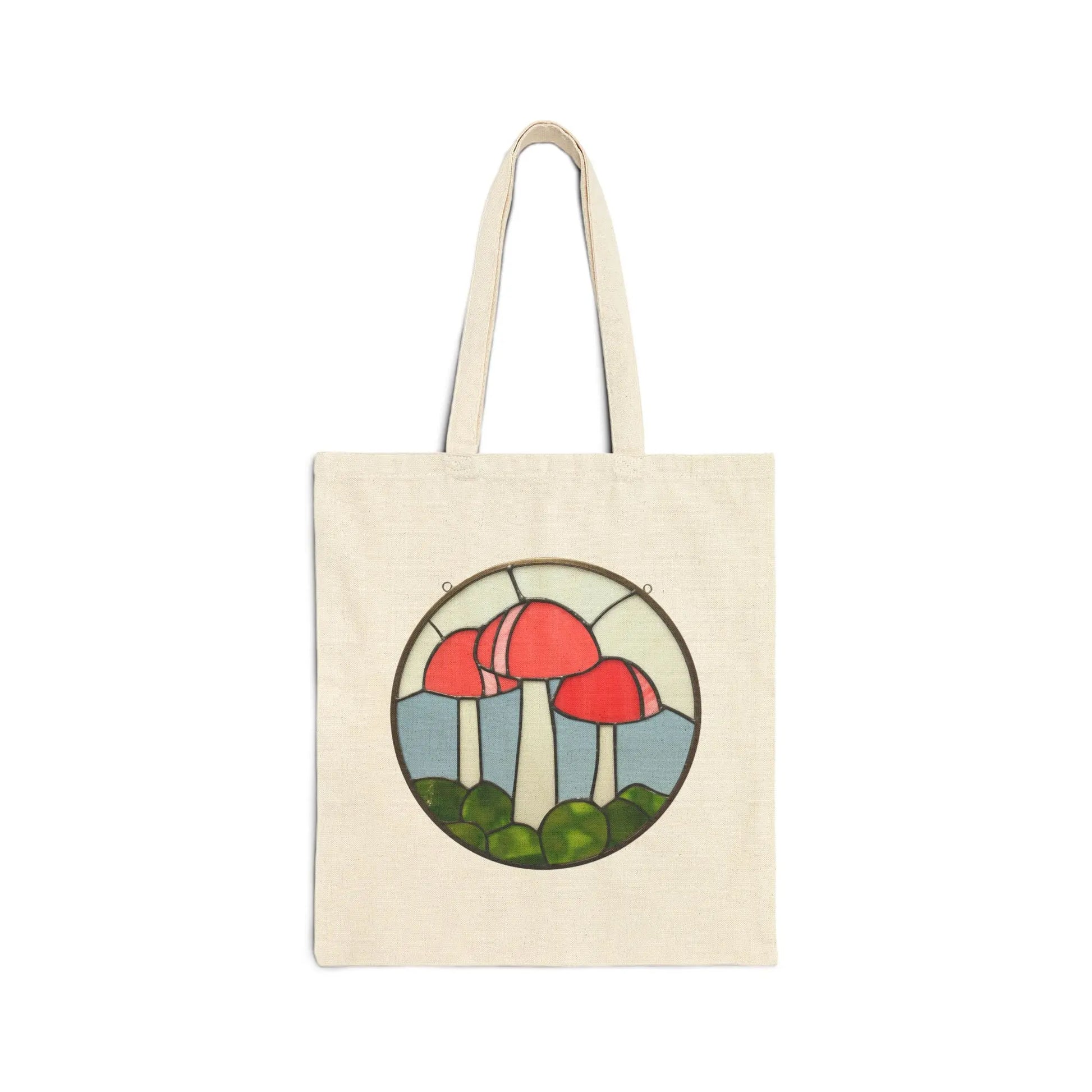 A natural cotton canvas tote bag by Noelle Barnes, named Stained Glass Mushrooms, showcases a circular design with three red mushrooms on white stalks and green grass at the base, resembling stained glass art.
