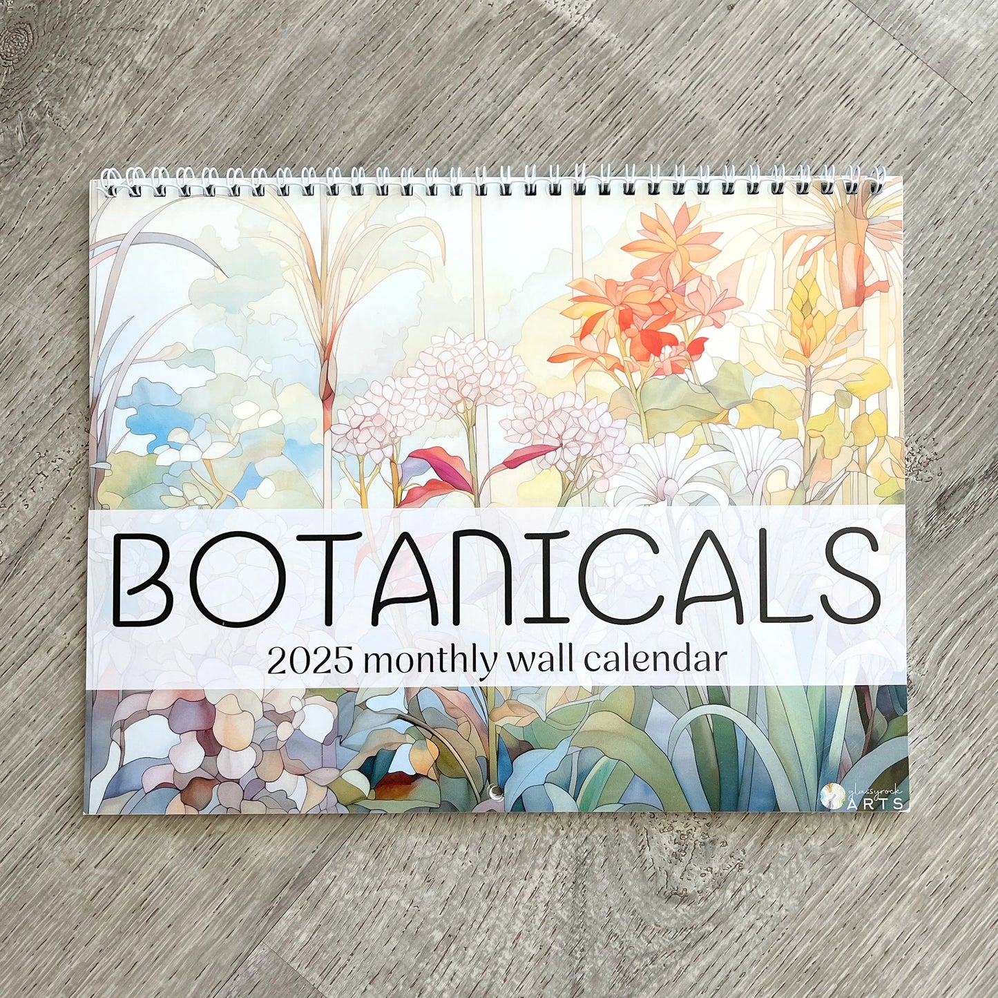The 2025 Botanicals Wall Calendar adorns a wooden surface, featuring stunning botanical illustrations. Its glossy, spiral-bound cover presents a vibrant array of flowers and plants reminiscent of stained glass beauty.