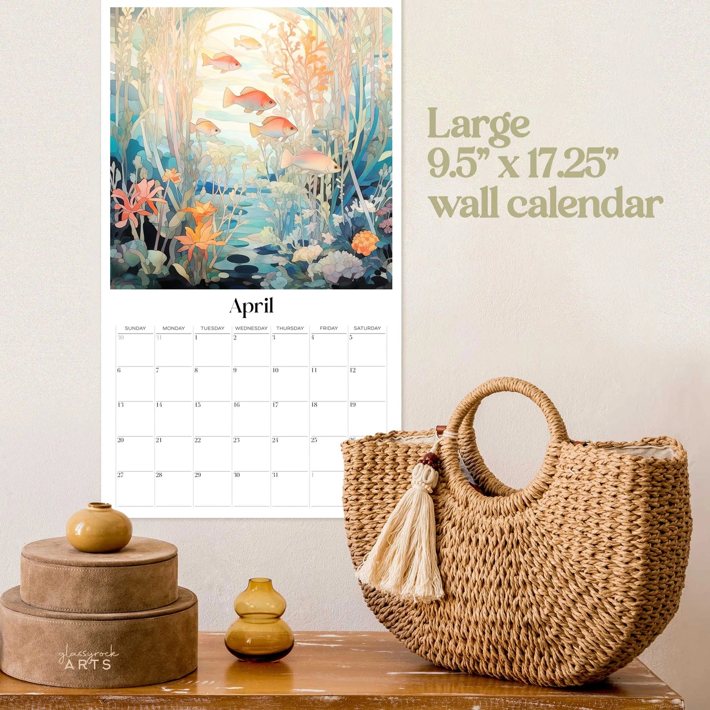 The 2025 Ocean Life Large Wall Calendar for April showcases fish and aquatic plants. Nearby, find a wicker bag with a tassel and stacked brown round boxes, complemented by a small yellow vase that adds charm to the ocean-themed art display.