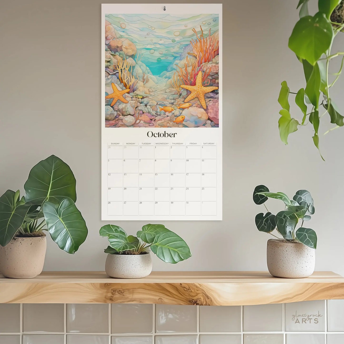 The 2025 Ocean Life Large Wall Calendar, showcasing a vibrant underwater October scene with starfish and coral, hangs above a wooden shelf. Three potted plants decorate the shelf, while a hanging plant adds charm on the right. The ocean-themed art is bright and colorful.