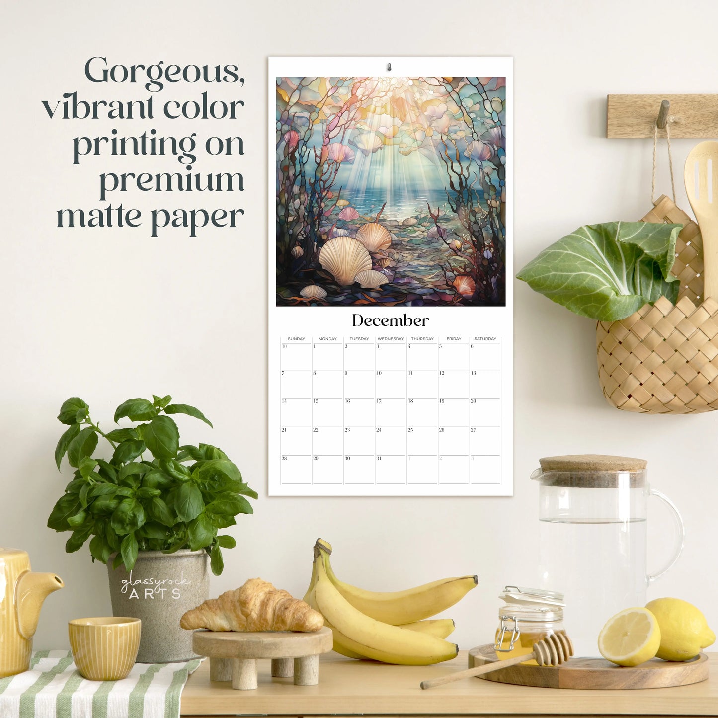 The 2025 Ocean Life Large Wall Calendar showcases a December page with seashells and sunlight, echoing ocean-themed art. Pictured above a kitchen counter with a wooden tray, glass pitcher, bananas, plant, and utensils. Features vibrant color printing on premium matte paper.