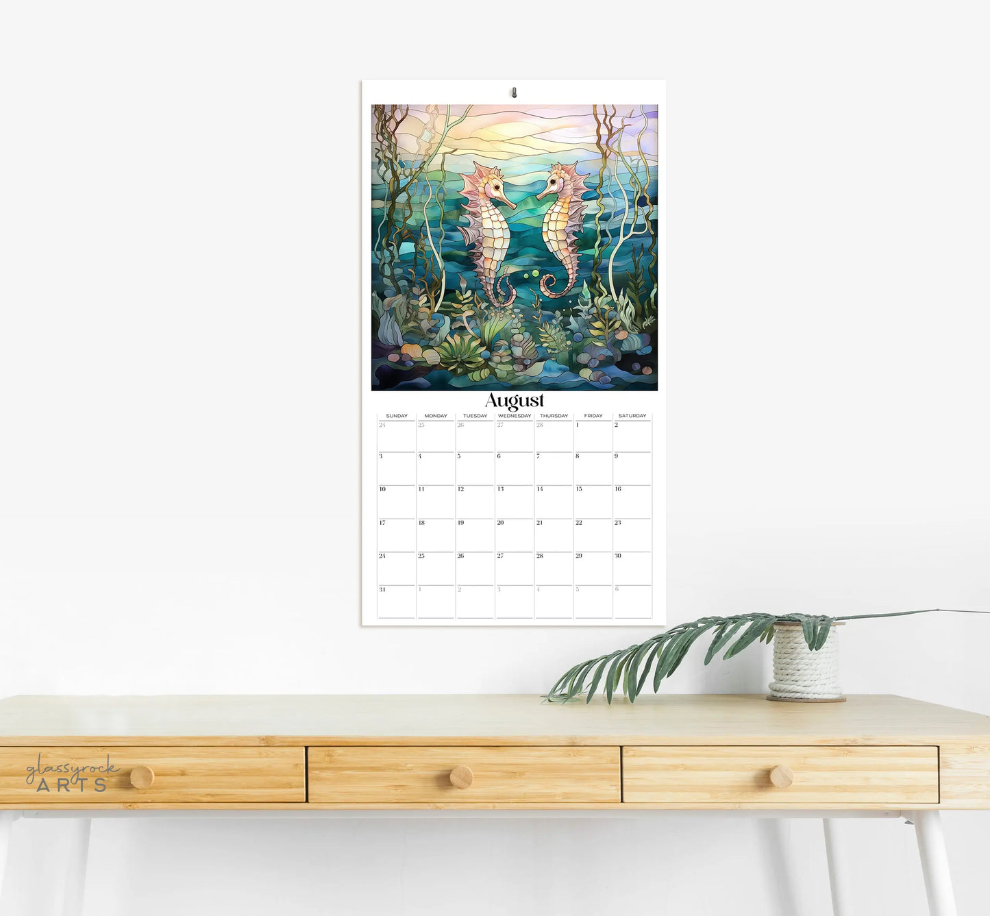 The 2025 Ocean Life Large Wall Calendar features an August page with illustrated seahorses in an underwater scene. It hangs above a light wooden desk, complemented by a potted plant on the right, in a minimalist room.