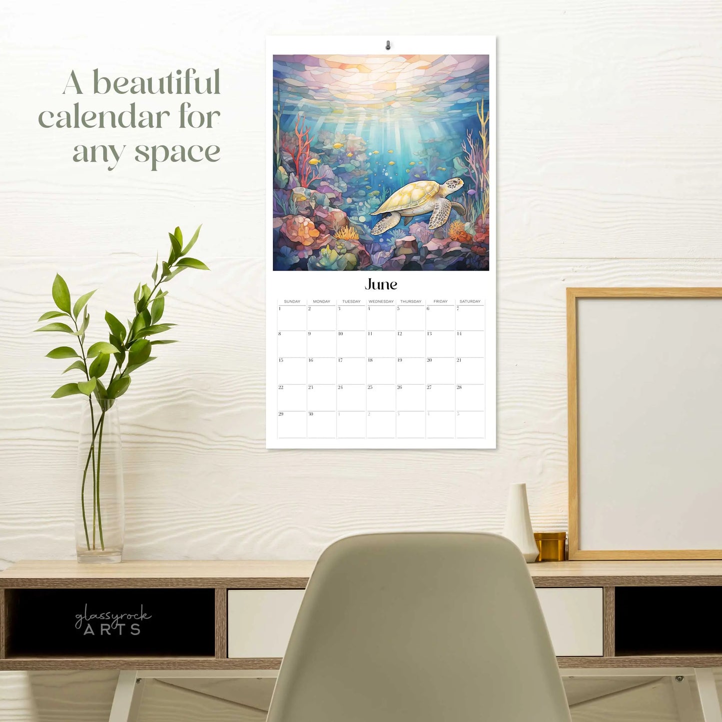 The 2025 Ocean Life Large Wall Calendar for June reveals a turtle swimming amidst vibrant coral under the sea. It hangs above a desk with a potted plant, framed art, and chair. Text reads, A beautiful calendar for any space.