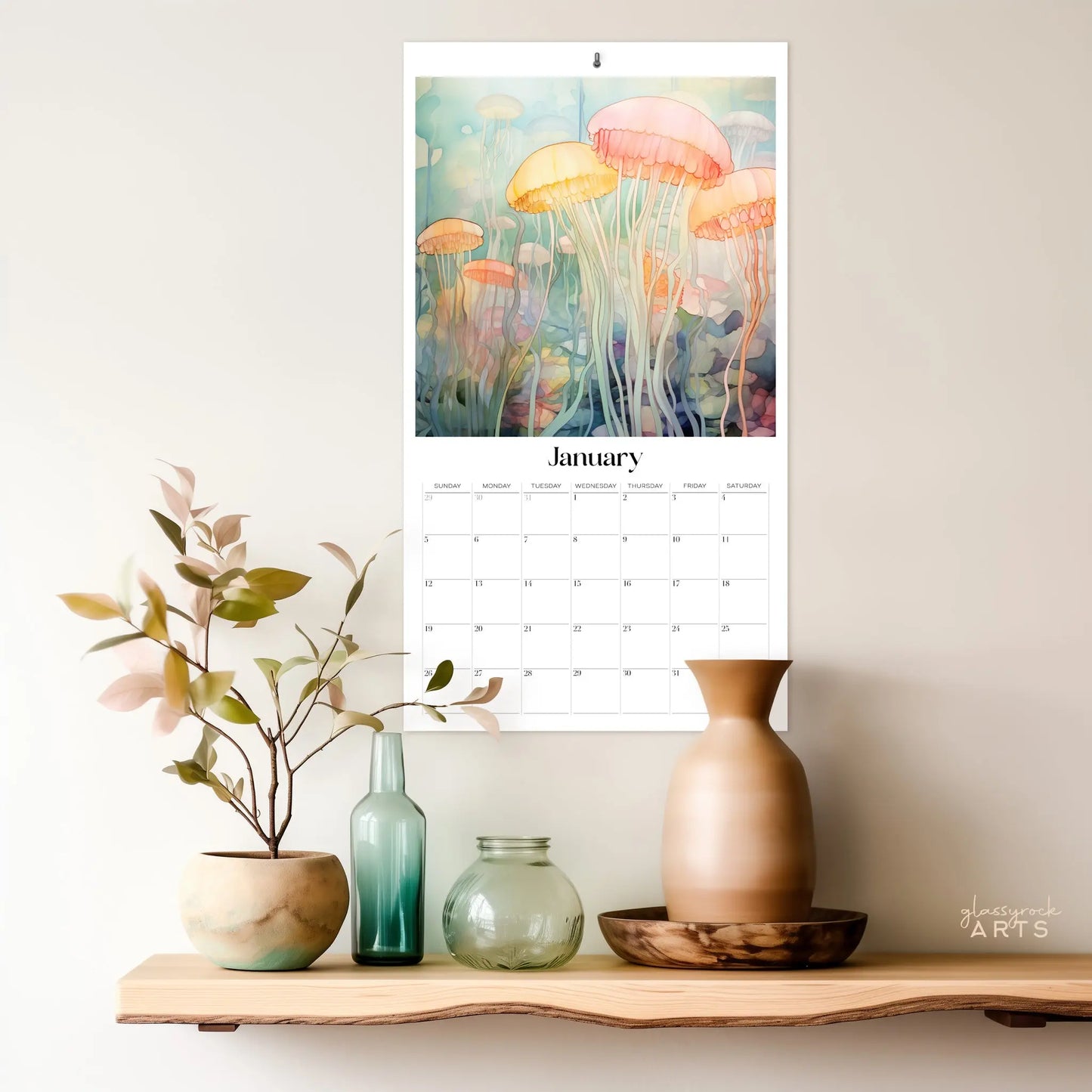 On a wooden shelf are decorative vases with plants, while above hangs the 2025 Ocean Life Large Wall Calendar displaying Januarys colorful, ethereal jellyfish in an underwater scene.