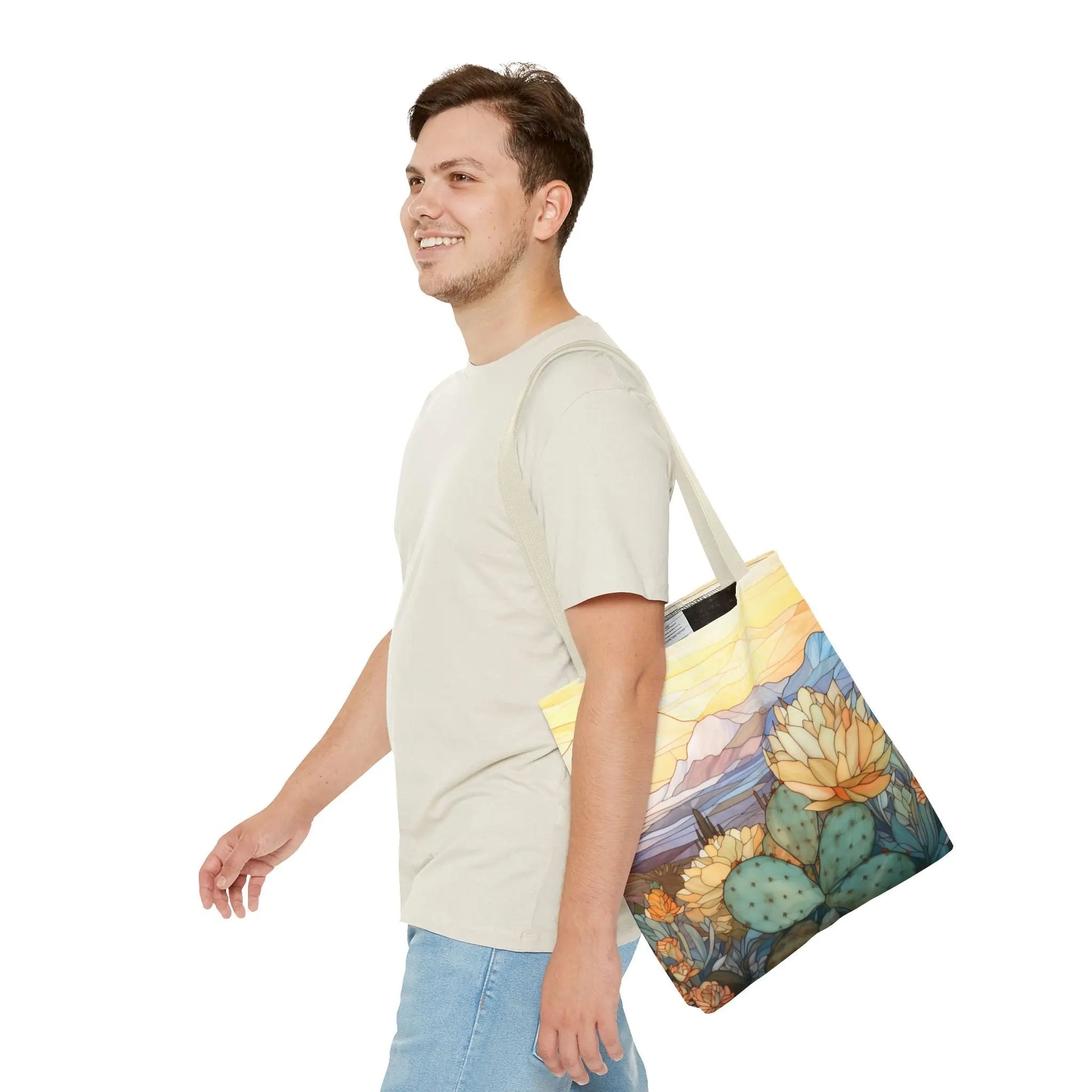 A person with short hair smiles, carrying a Stained Glass Desert Cactus Tote Bag featuring an artistic desert landscape. Dressed in a light t-shirt and jeans, they enjoy the durable polyester material that offers both style and longevity.