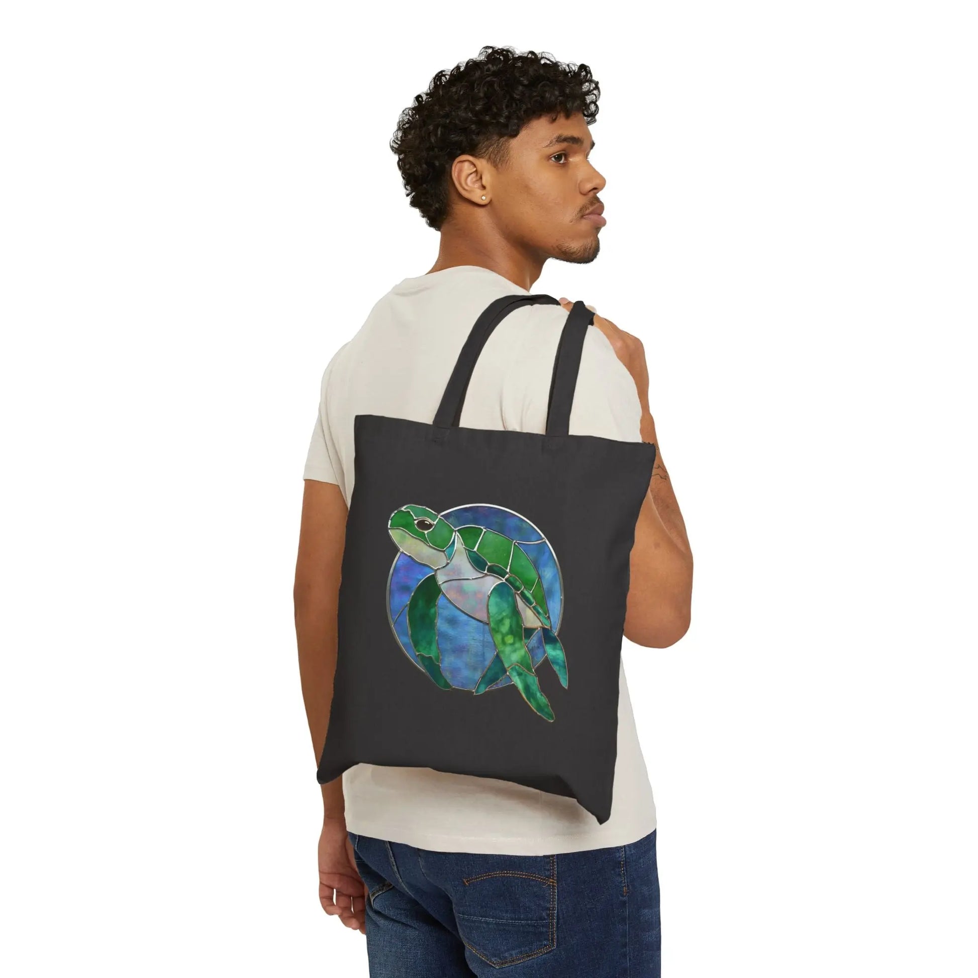 A person in a white shirt and jeans carries the Stained Glass Turtle Cotton Canvas Tote Bag over their shoulder, featuring a colorful sea turtle motif on plain white. Designed by Noelle Barnes.