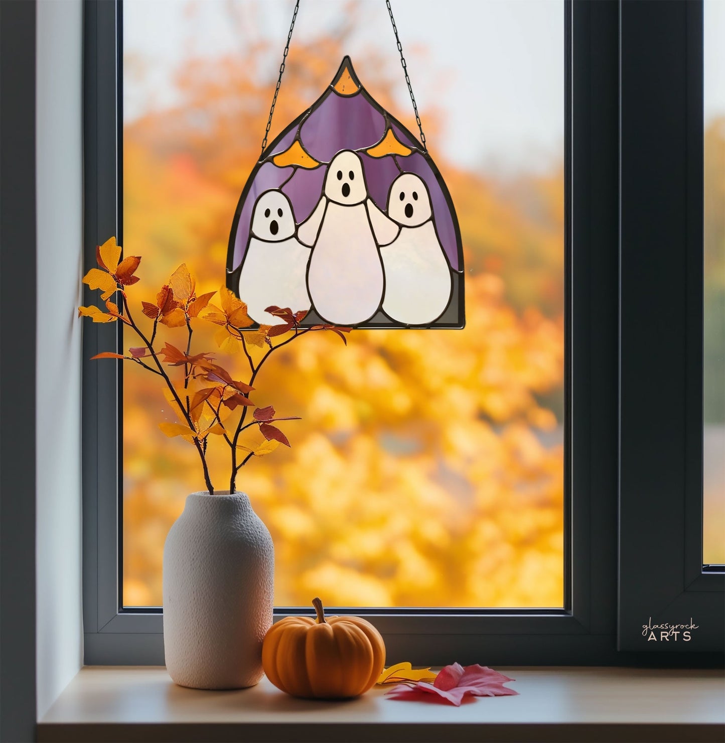 The Three Ghosts Halloween Stained Glass Pattern adorns a window, with autumn leaves blurred beyond and a white vase with branches and an orange pumpkin on the sill, creating an ideal scene for any stained glass enthusiast to draw inspiration from.